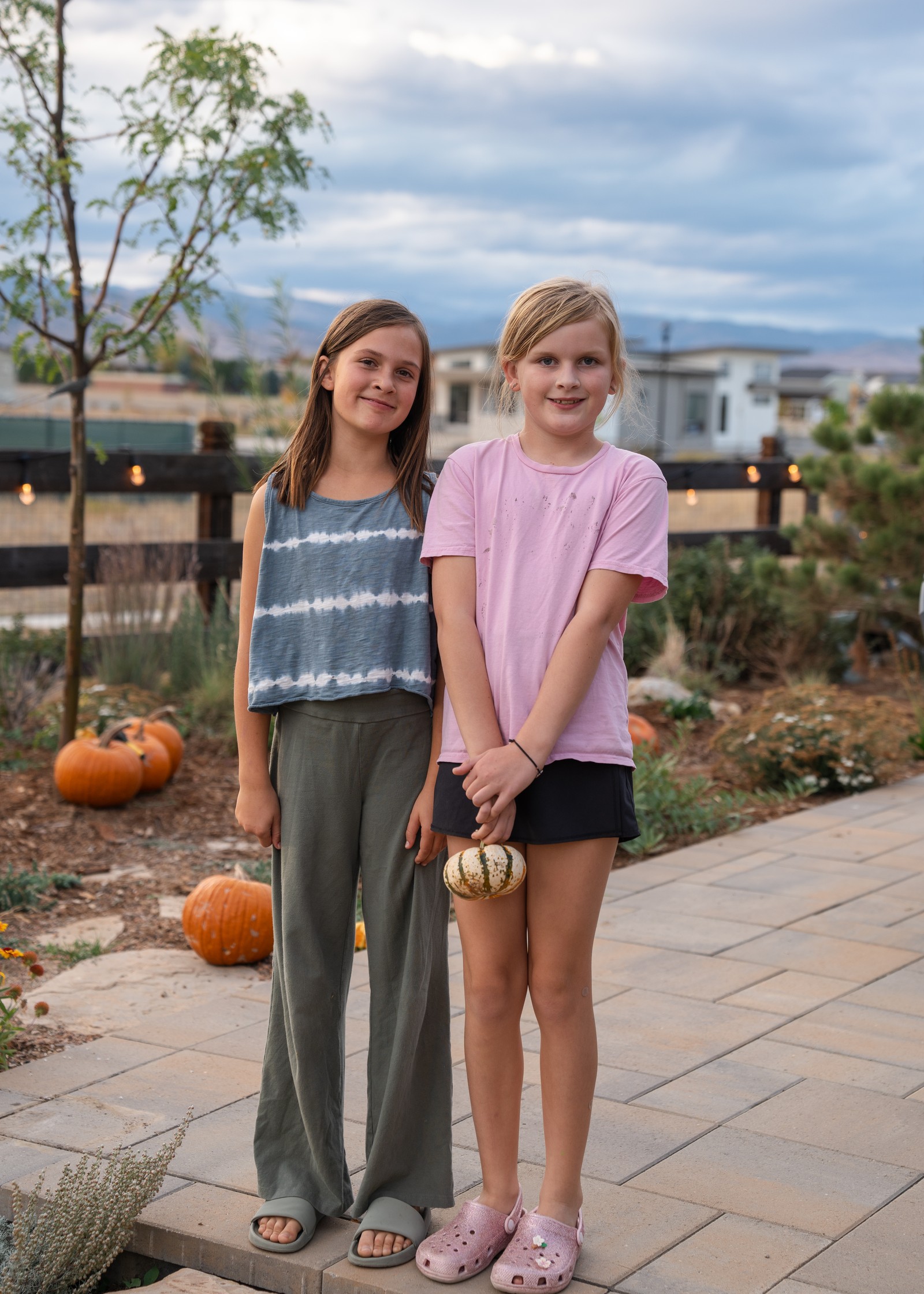 Two people stand side by side on a paver walkway, one in a gray striped top and one in a pink top, with pumpkins nearby.