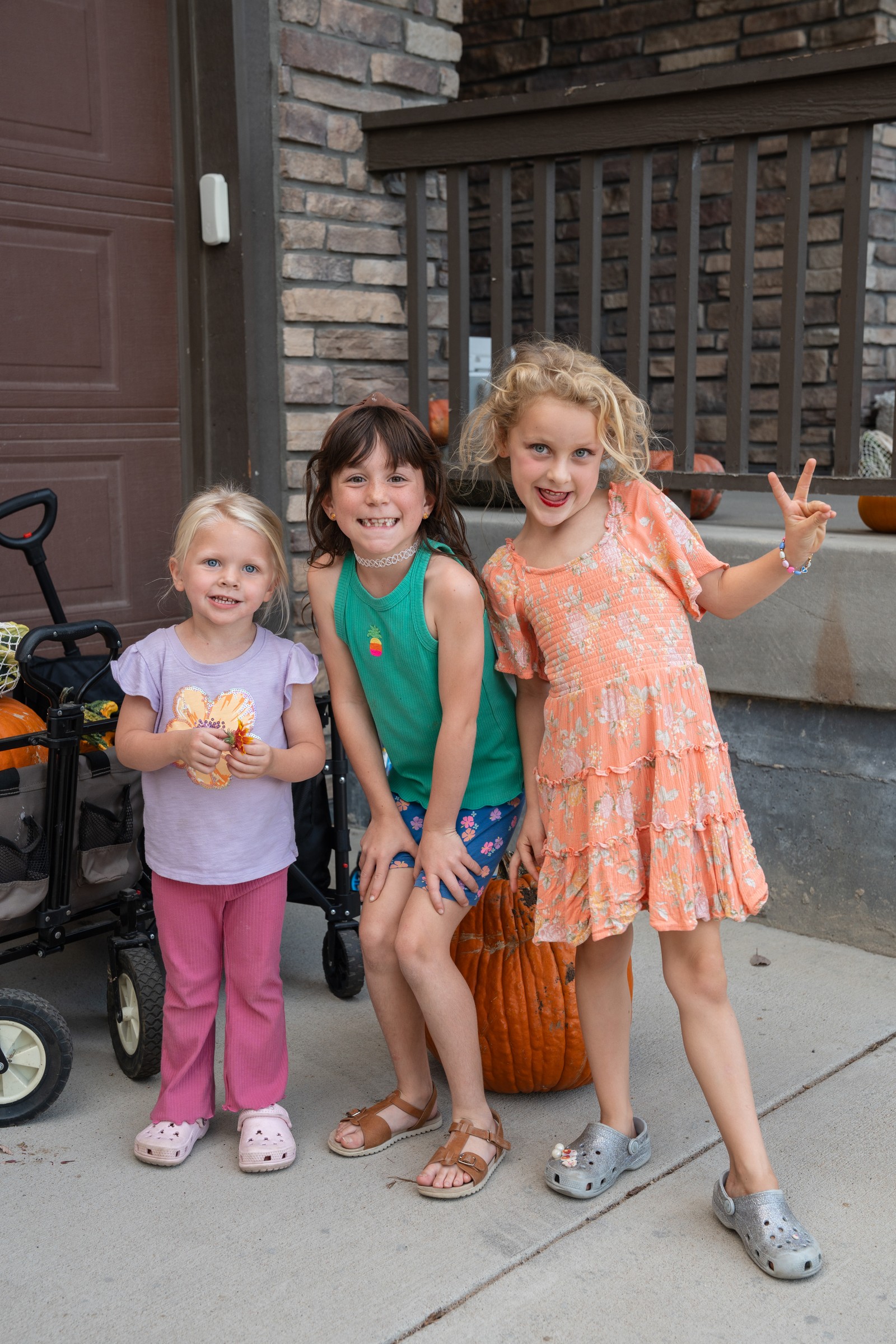 Three people stand on a patio in front of a stone wall, in light pink, teal and orange outfits, with a stroller behind them.