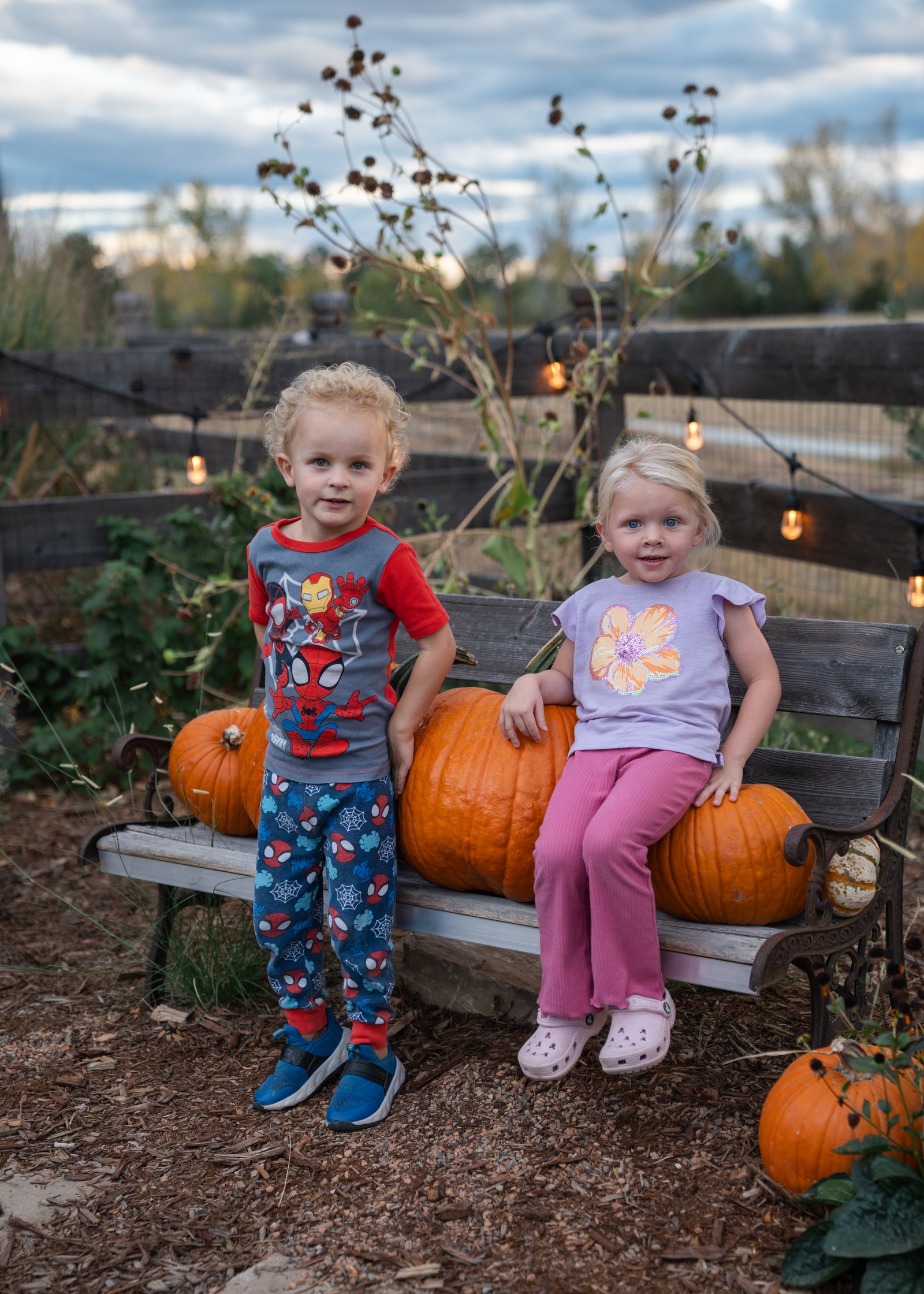 Two people sit on a garden bench with pumpkins, one in blue patterned pants and one in pink pants.