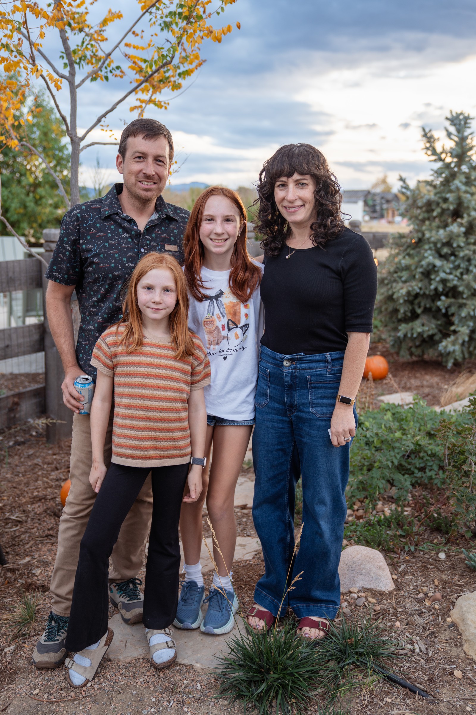 Four people stand together on a garden path at dusk, smiling at the camera, with autumn trees behind them.