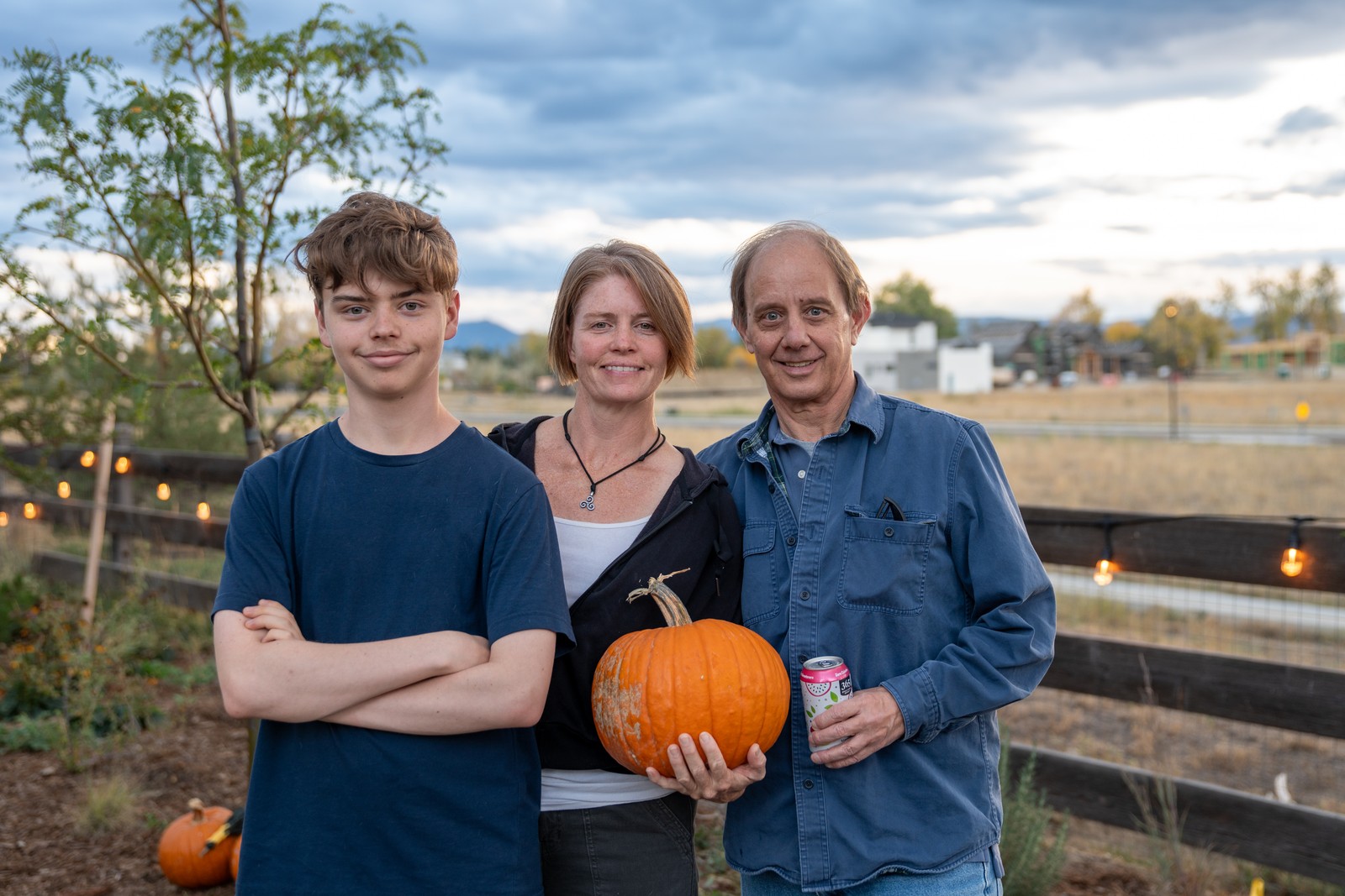 Three people stand together smiling, with the one at right holding a pumpkin, in front of string lights and a rail fence.