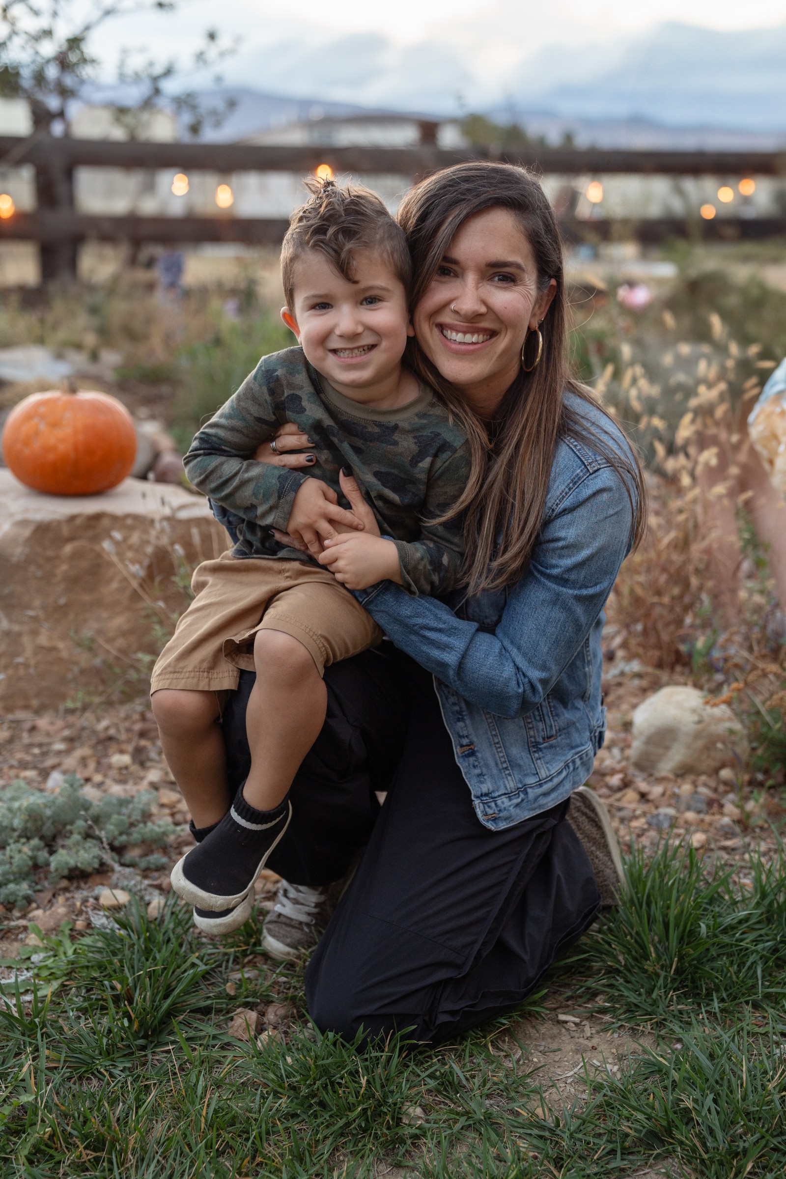 A person in a denim jacket kneels in a garden bed and smiles while holding someone in a camouflage-print top, with a pumpkin on a boulder behind them.