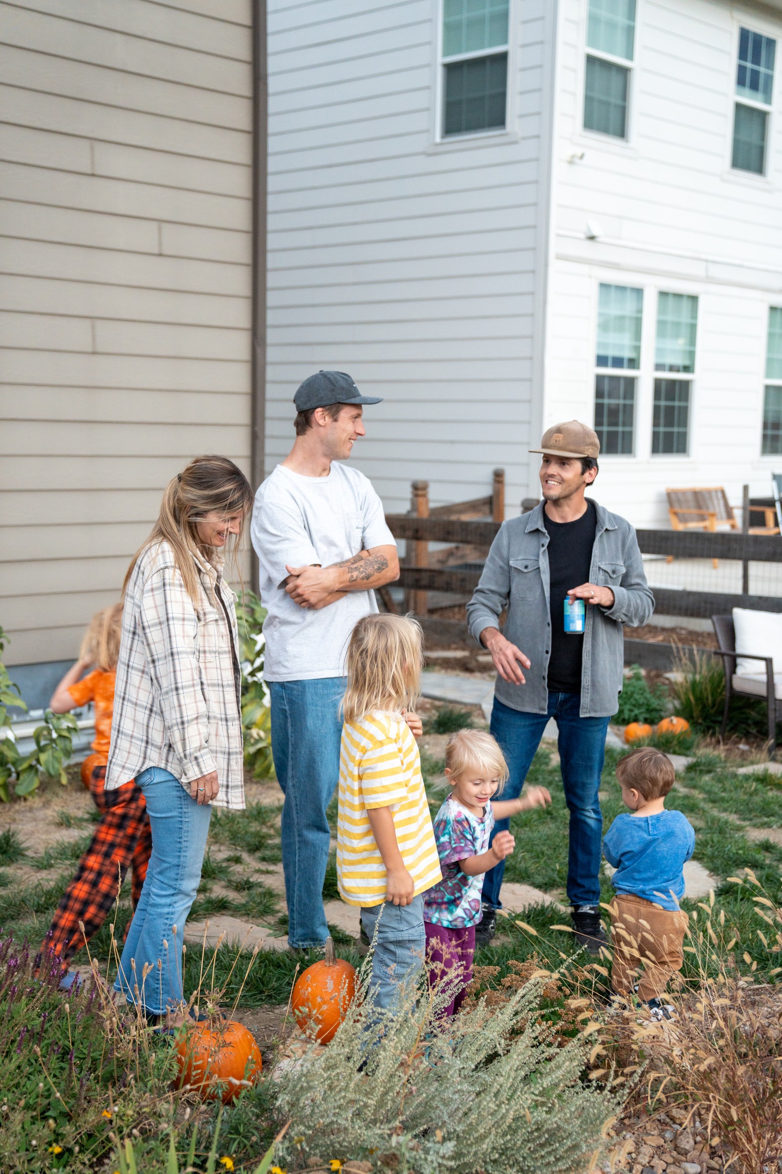 Guests stand and talk in a yard among pumpkins, in front of two houses with light siding.