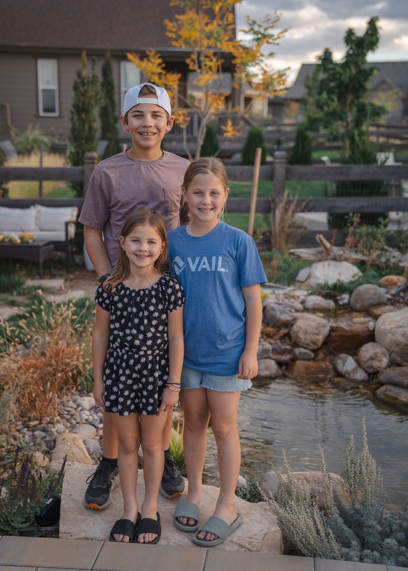 Four people stand together beside a pond, in blue, gray and dark patterned outfits, with houses behind them.