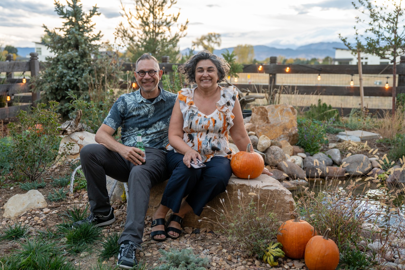 Two people sit together on a boulder in a garden with pumpkins at their feet and hazy hills behind them.