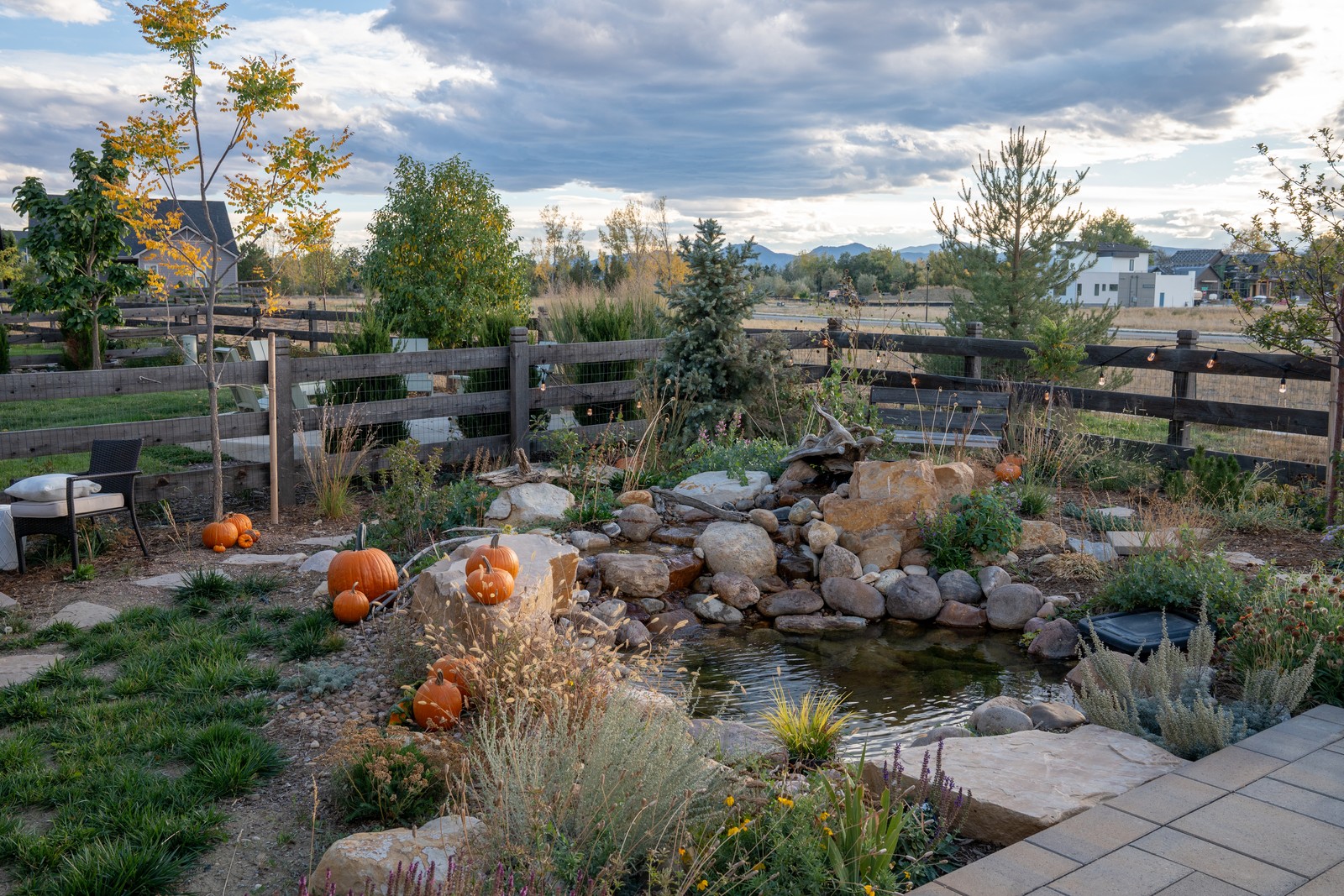 The edge of a boulder-lined pond with pumpkins nearby and a paver patio corner at the lower right.