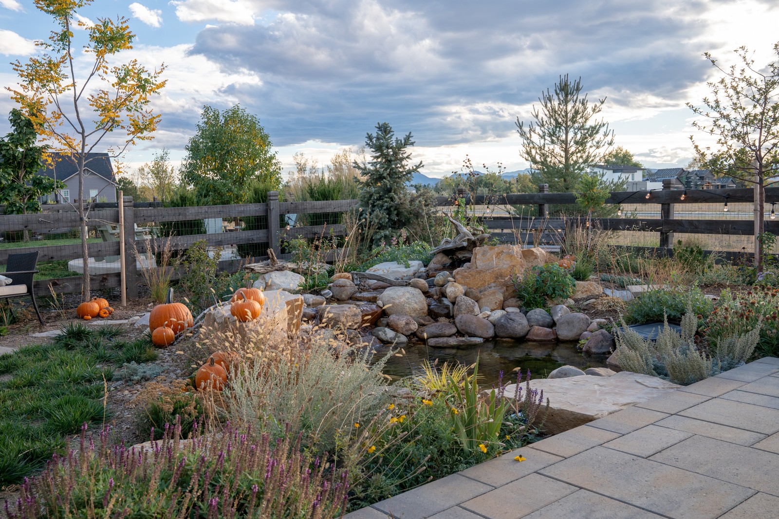 An evening sky arches over a paver patio, a boulder-edged pond and pumpkins in a landscaped backyard.