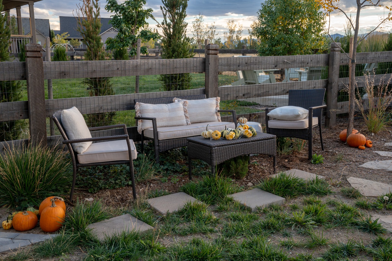An outdoor seating area with cushioned wicker chairs, a sofa and a low table with small gourds, framed by a wooden fence and pumpkins on the lawn.