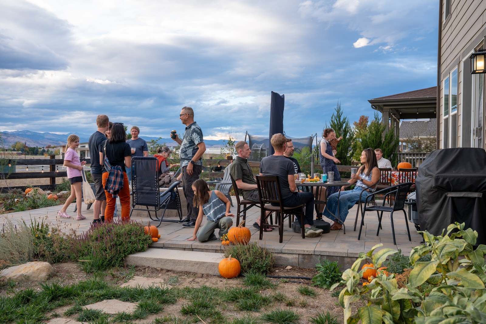 Guests gather at outdoor tables on a patio at dusk, with a pond-side garden and a house at the right edge.
