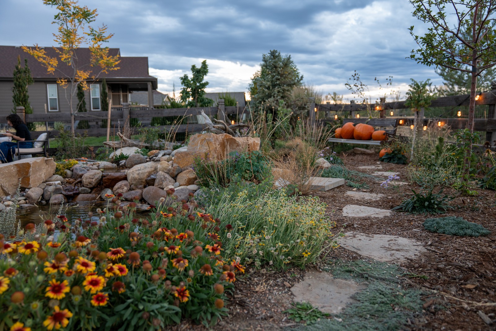 Orange and yellow blanket flowers bloom along a stepping-stone path in a backyard garden, with string lights and a bench of pumpkins in the distance.
