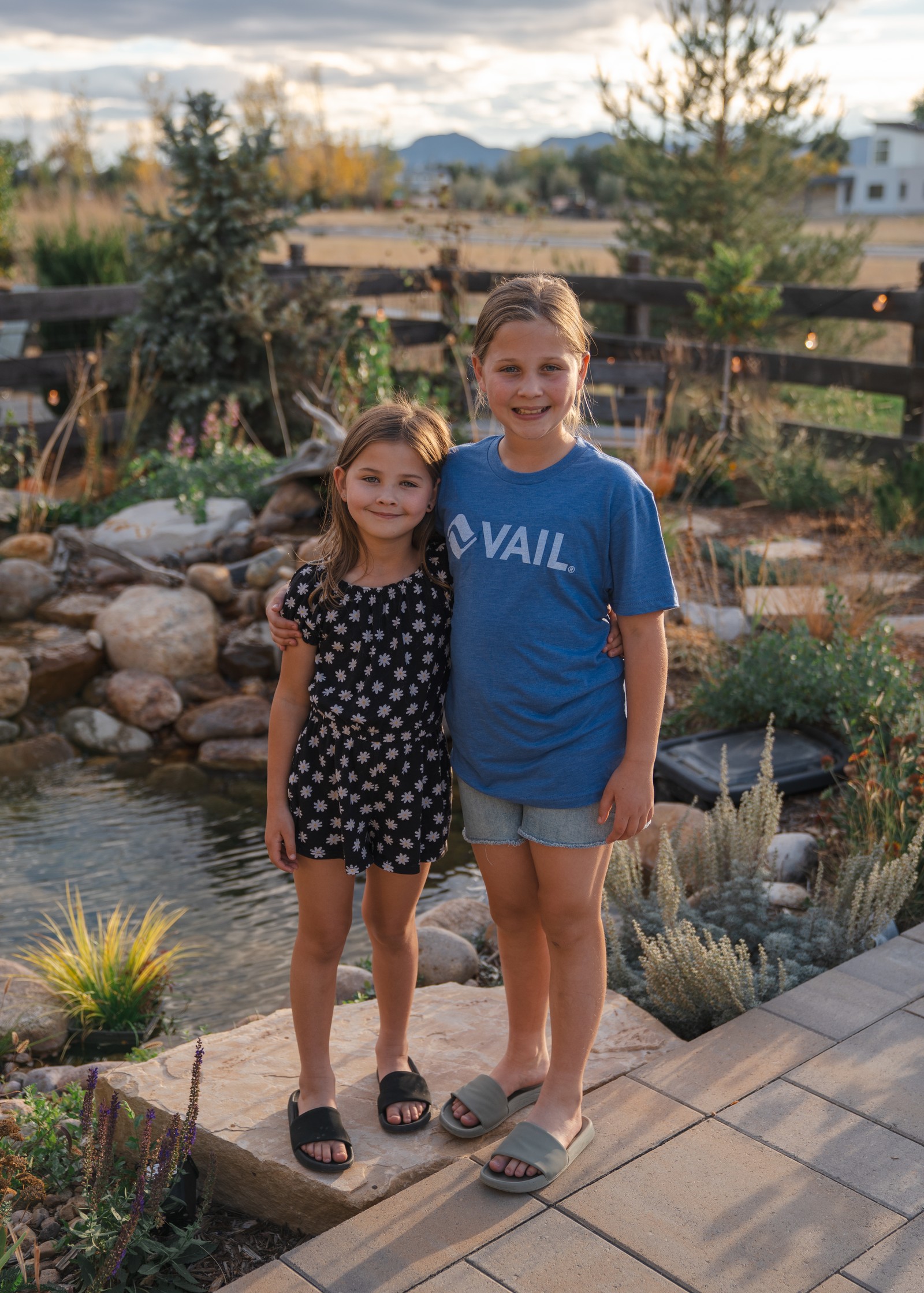 Two people stand side by side on a paver landing beside a boulder-edged pond, one in a blue t-shirt and one in a dark patterned dress.