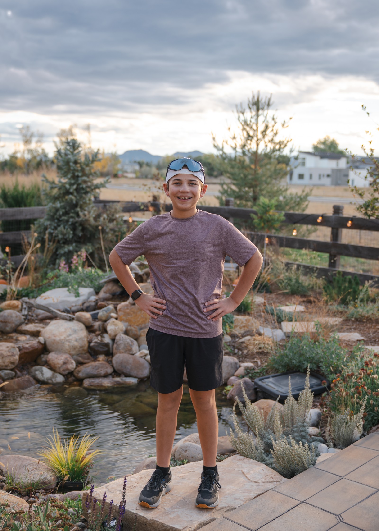 A person in a gray t-shirt and dark shorts stands on a stone landing beside a backyard pond at dusk.