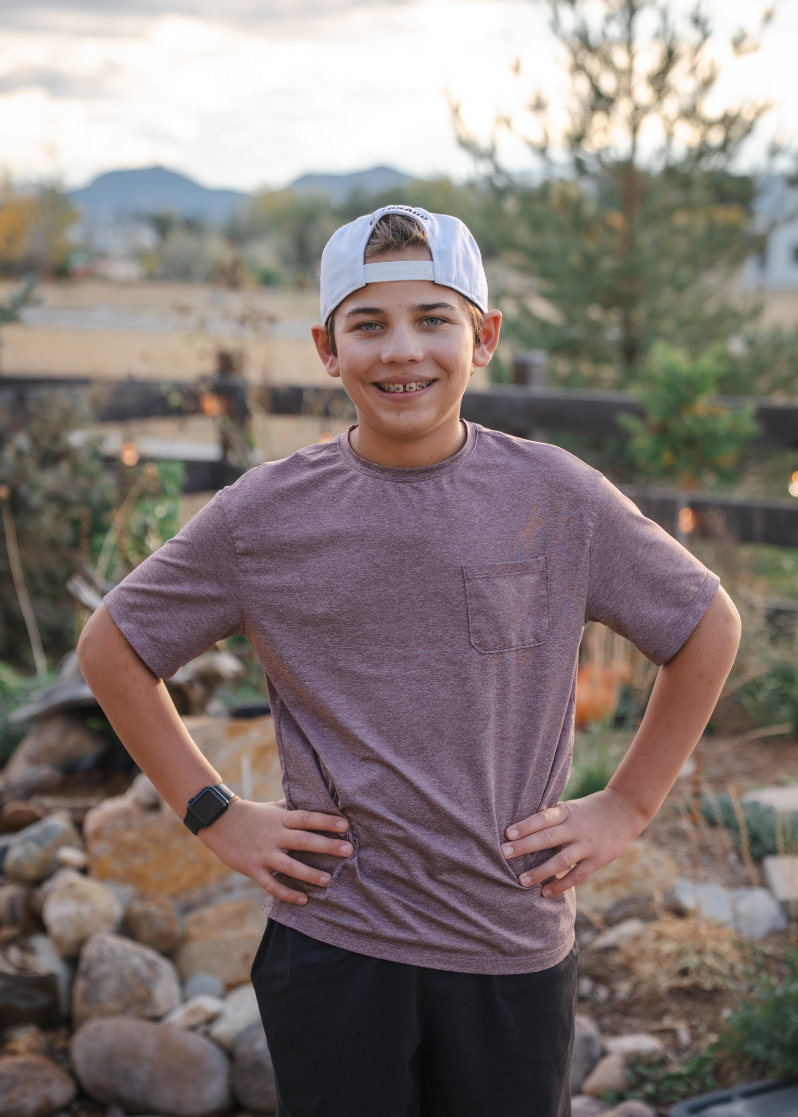 A person in a baseball cap and gray t-shirt stands smiling with hands on hips in a rock-lined backyard garden.