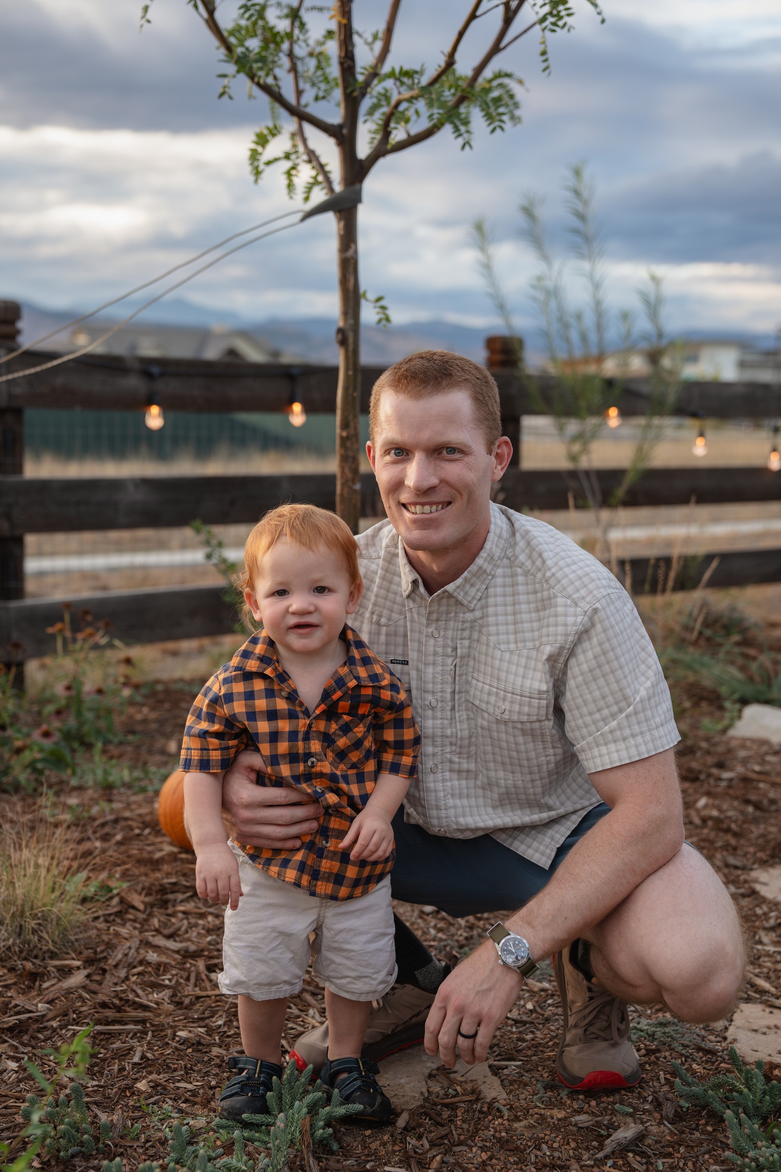 Two people pose in a backyard garden in front of a wooden fence, one in a plaid shirt and one in a light button-up shirt.