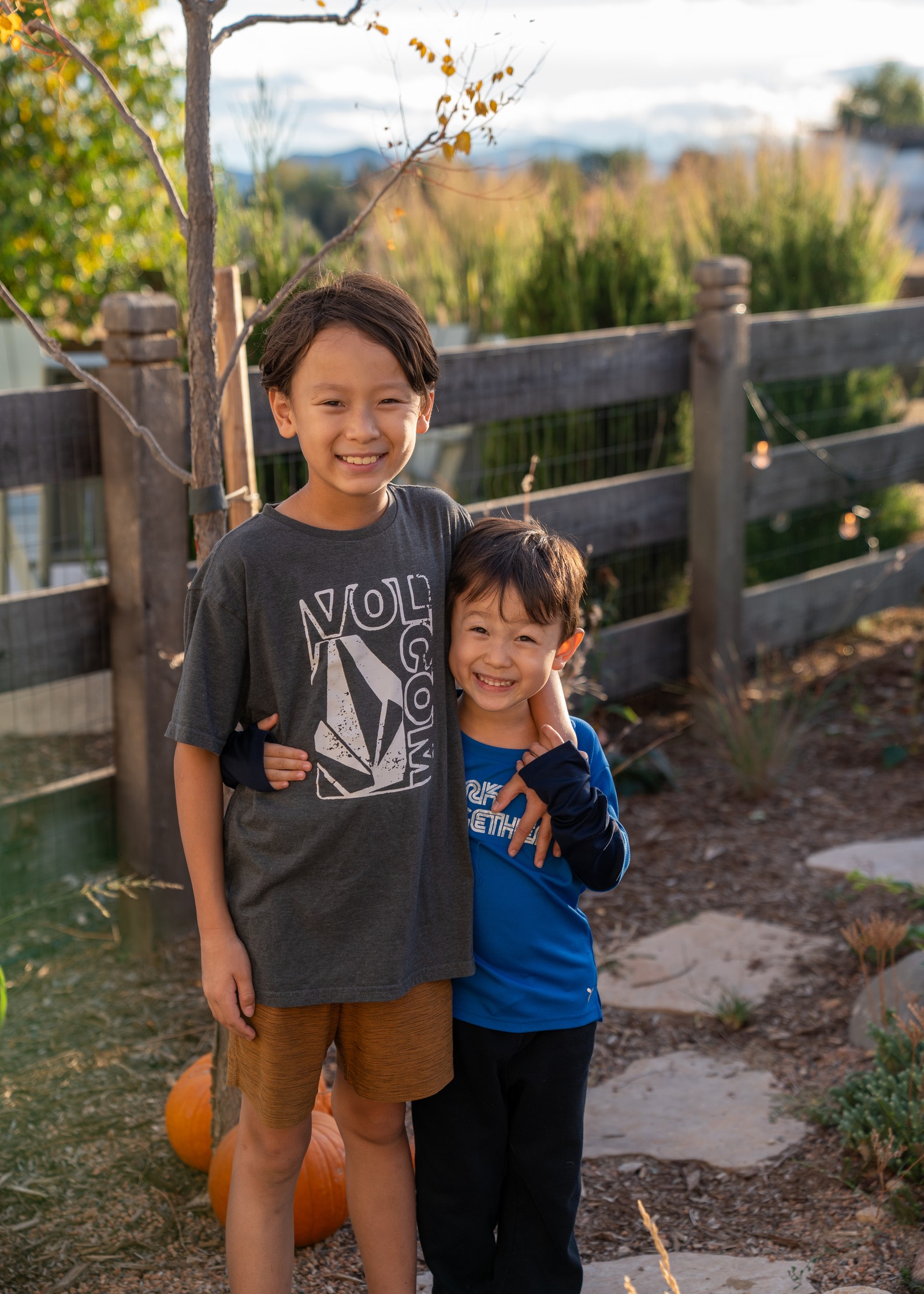 Two people stand side by side in front of a wooden fence, one in a gray graphic t-shirt and one in a blue shirt, smiling.