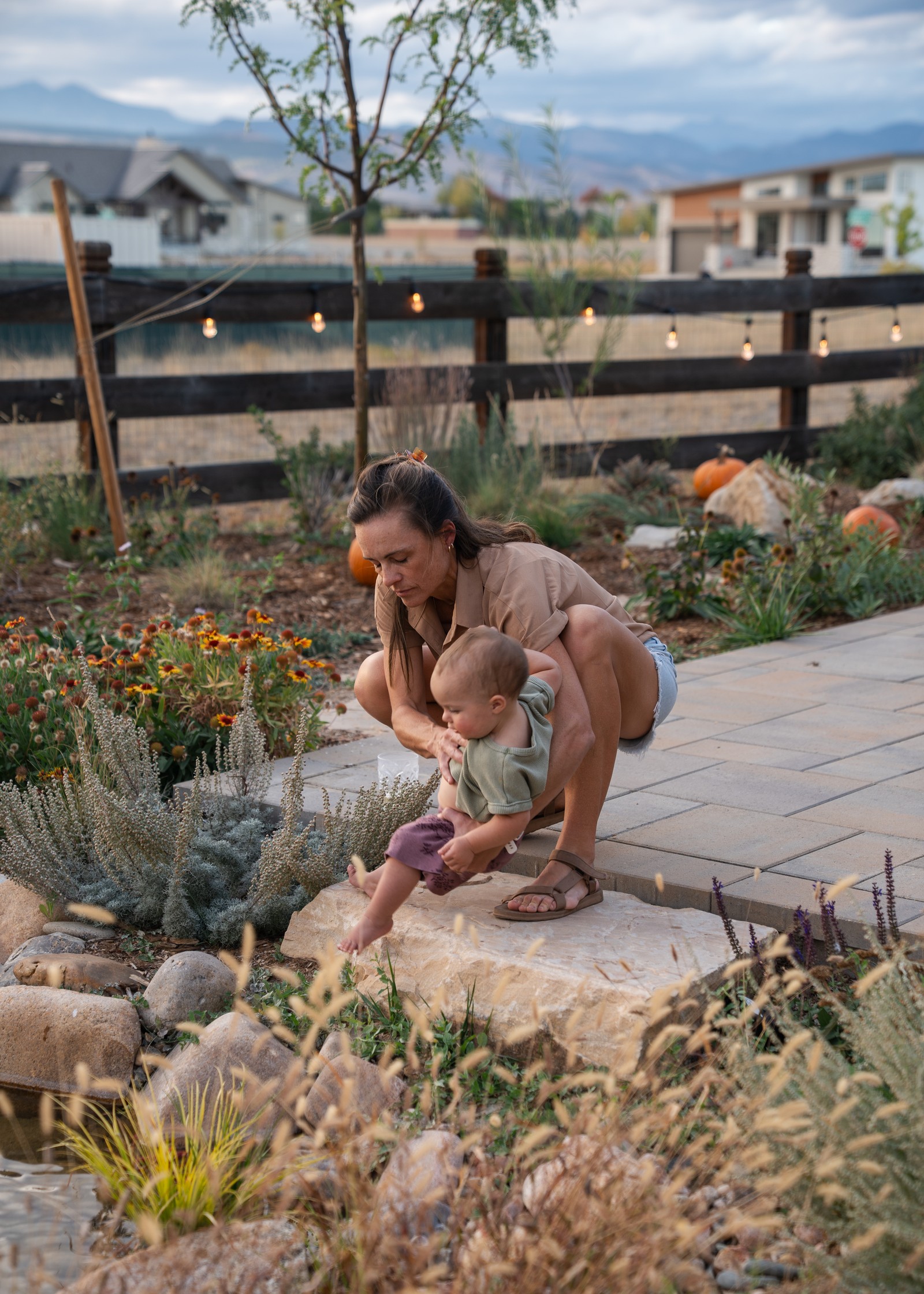 A person crouches at the edge of a flat stone path in a garden with grasses and flowering plants, looking down.