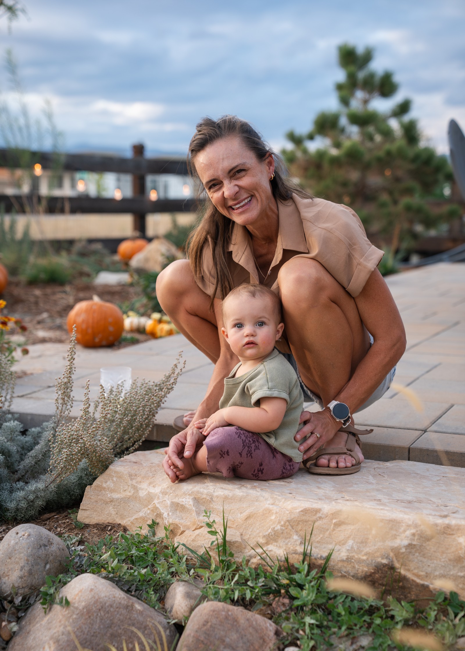 Two people sit on a stone ledge in a garden, smiling at the camera, with pumpkins and evergreen trees behind them.