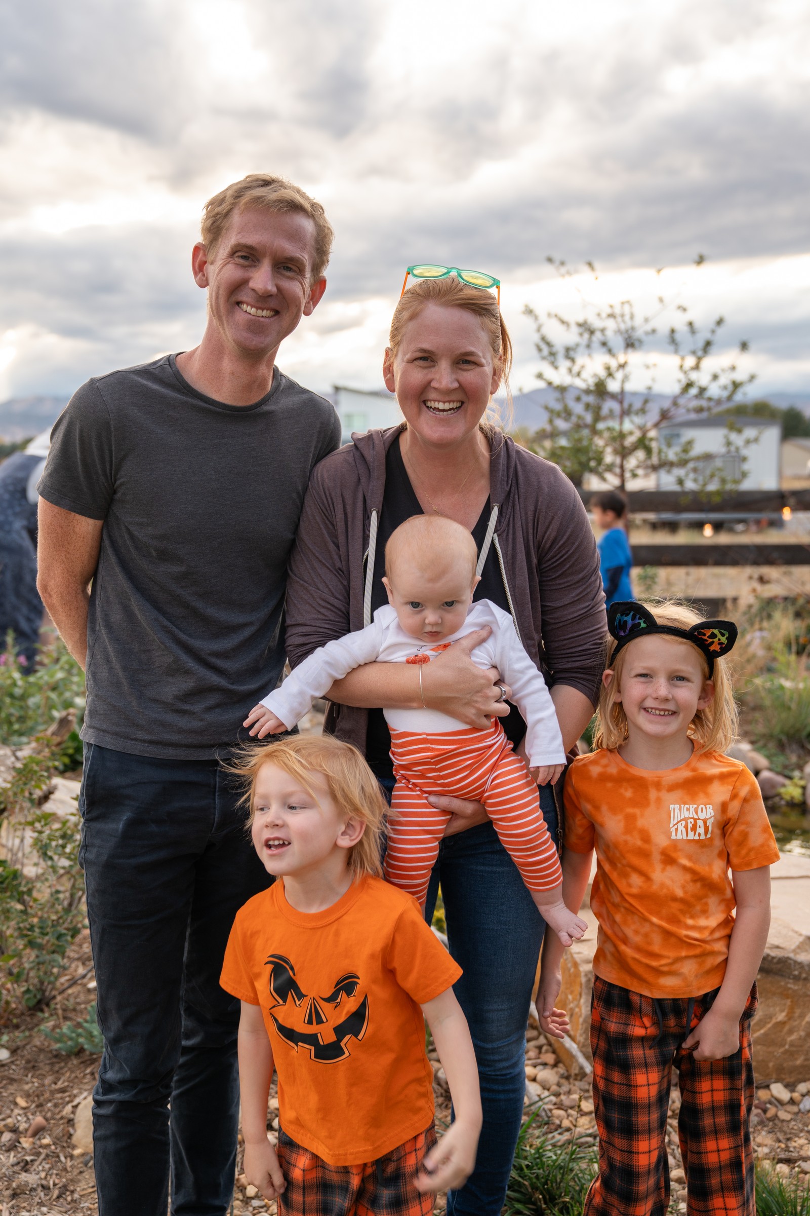 A group poses in a yard at dusk, with two people in matching orange pumpkin shirts and plaid pants.