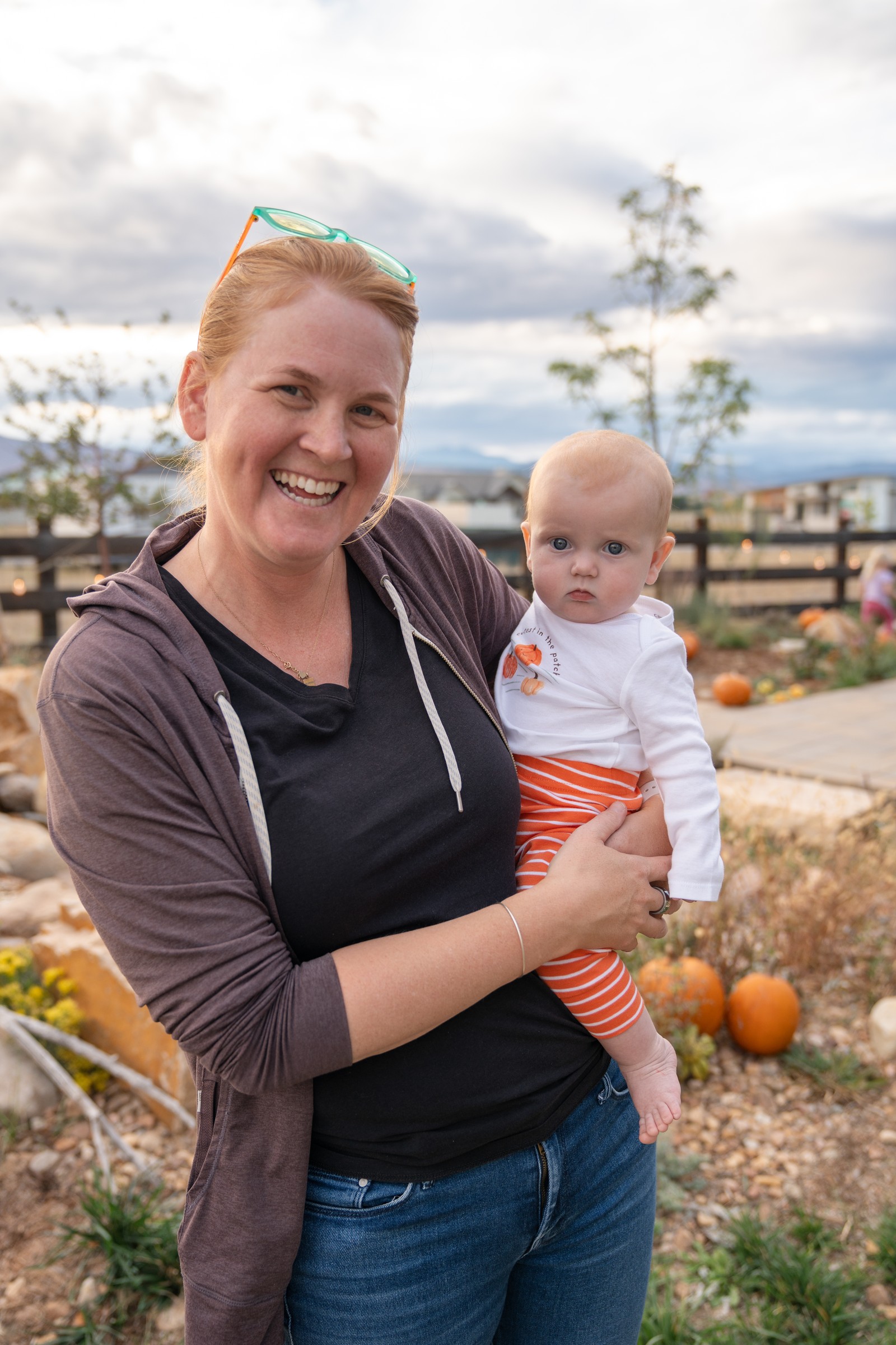 A person in a dark top smiles while holding someone in a white top and orange pants, with a yard and fence behind them.