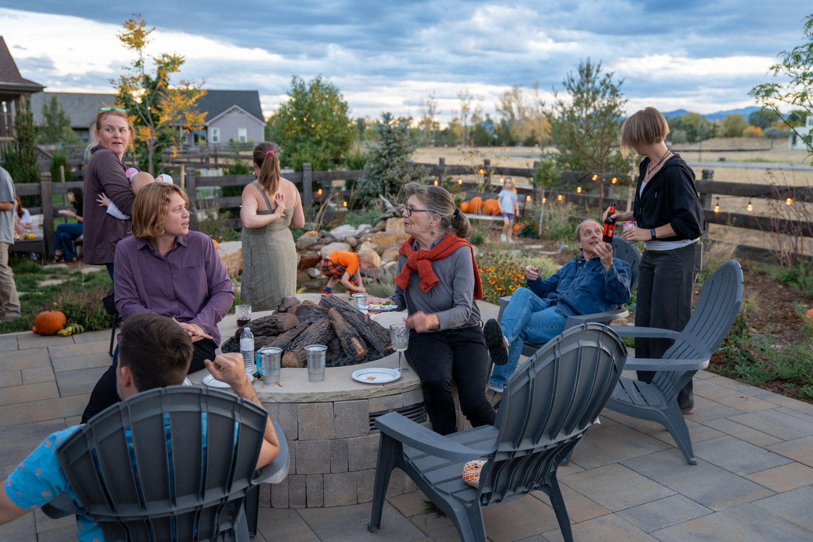 Guests sit and stand around a stone fire pit table on a patio at dusk, with outdoor chairs in the foreground.