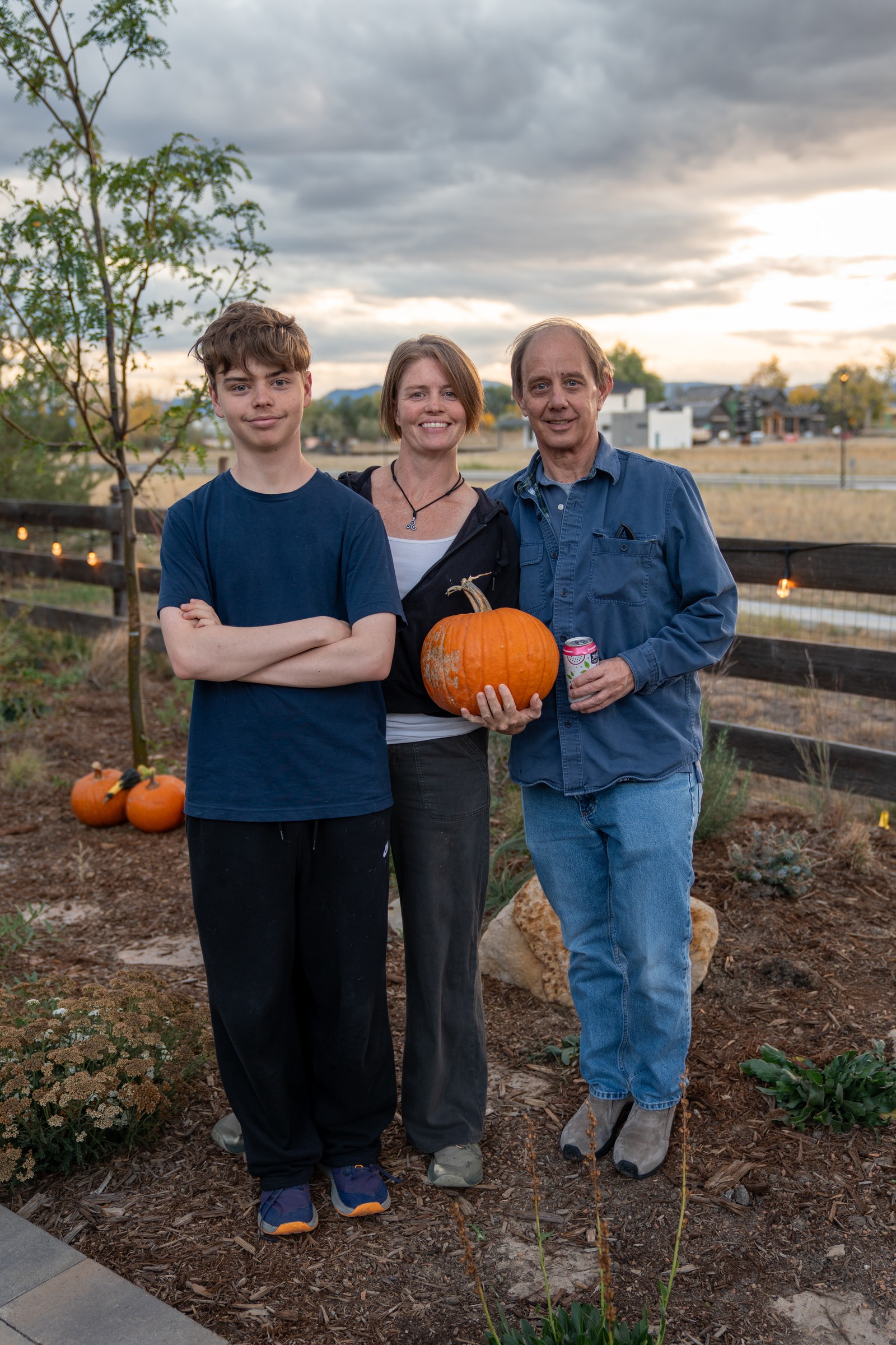 A small group stands together in a yard at dusk, with one person holding a pumpkin, in front of a wooden fence.