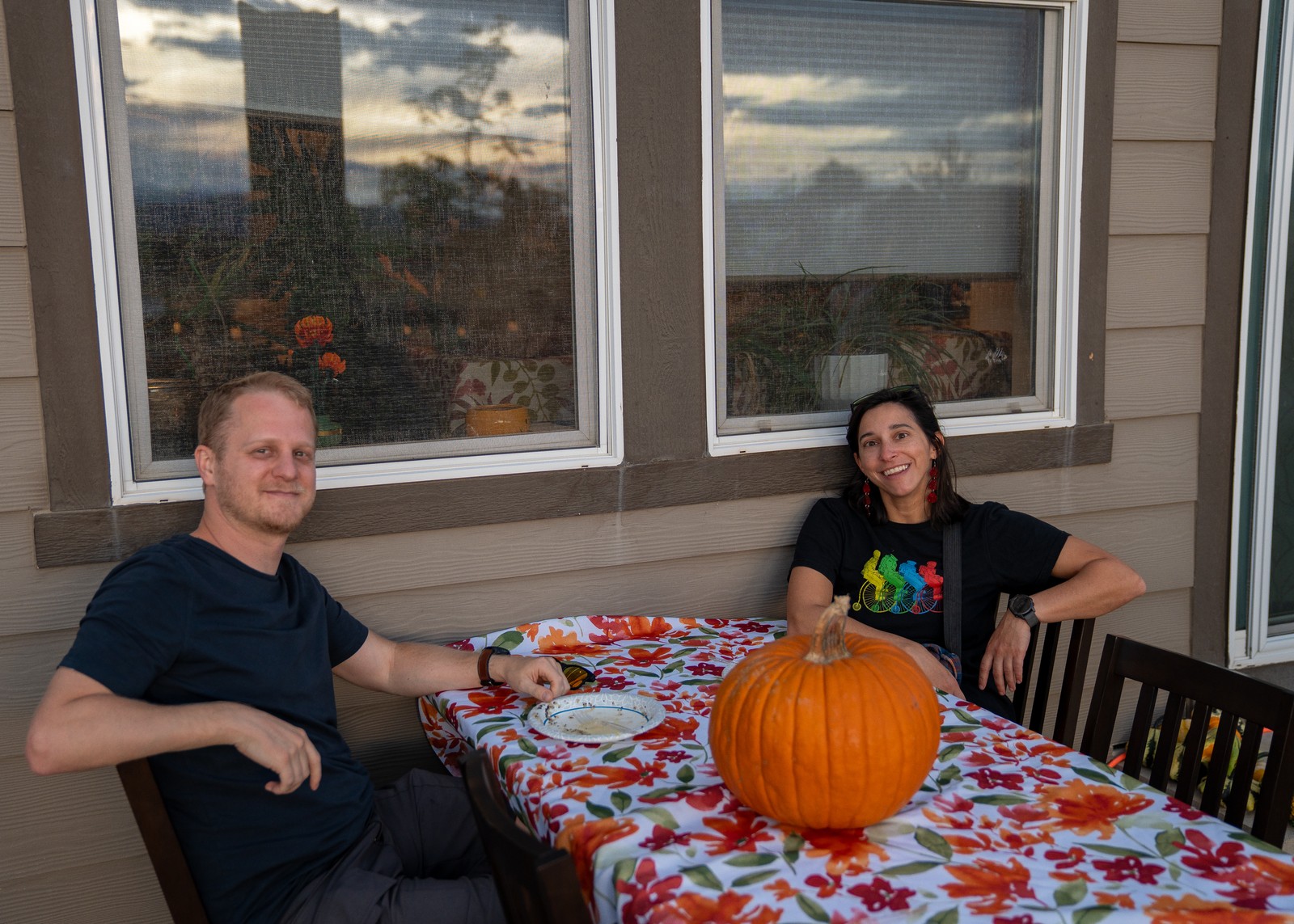 Two people sit at a table covered in a bright floral cloth with a large pumpkin, in front of large windows.