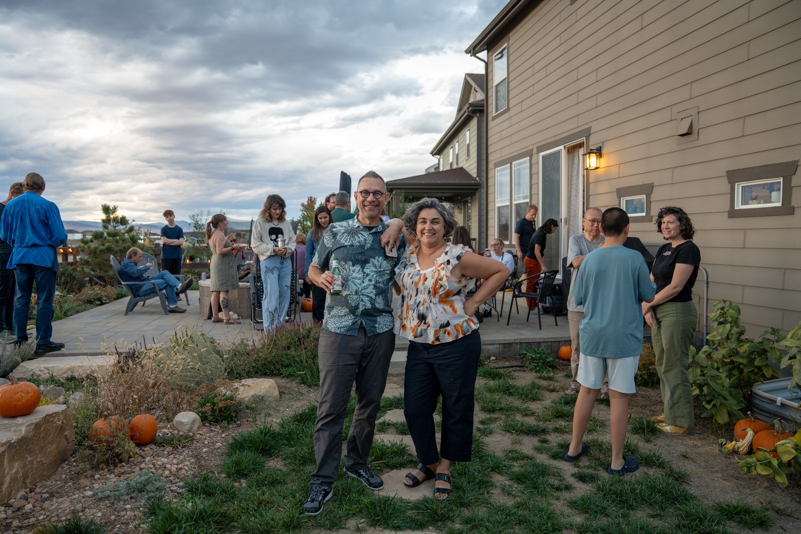 Guests mingle on a patio beside a house at dusk, with two people smiling in the foreground.