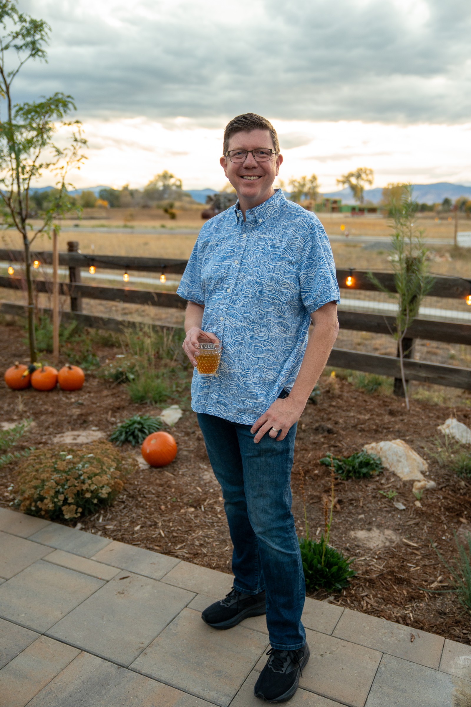 A person in a light blue patterned shirt stands on a paver path in a garden with pumpkins and a wooden fence.