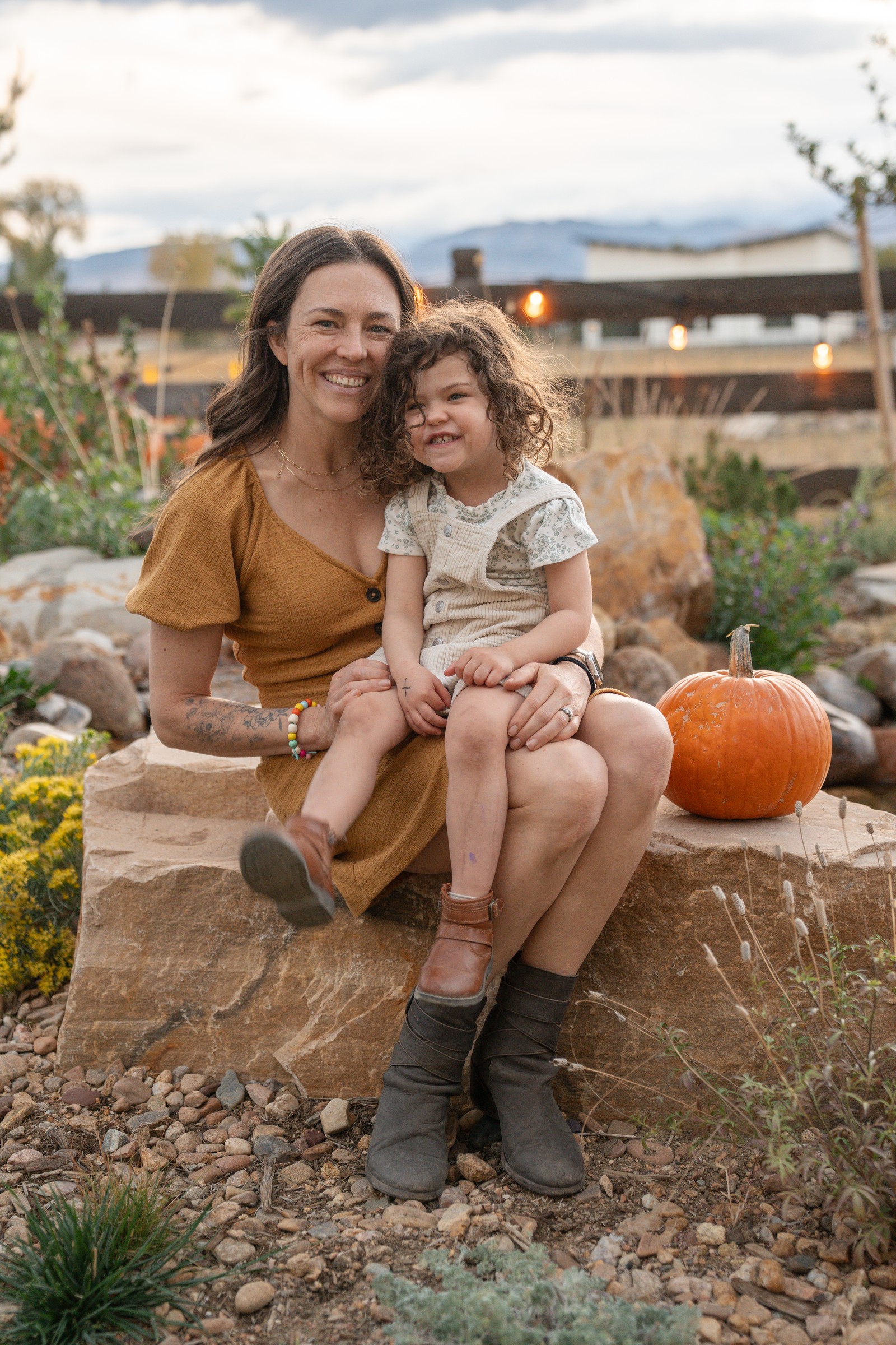 Two people sit on a boulder ledge next to pumpkins, one in a mustard dress, smiling at the camera.