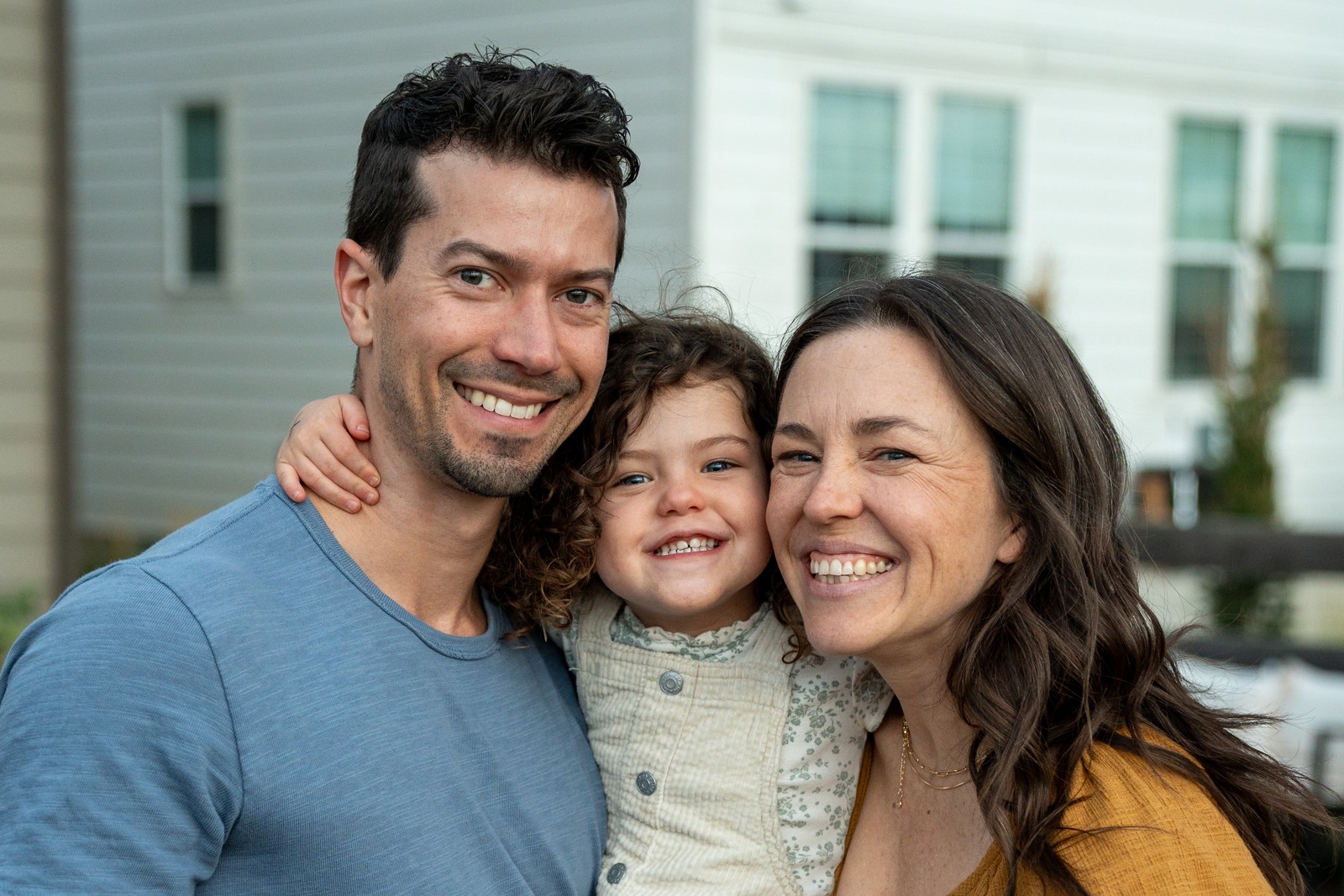 Three guests smile cheek to cheek, with a light-colored house softly blurred behind them.