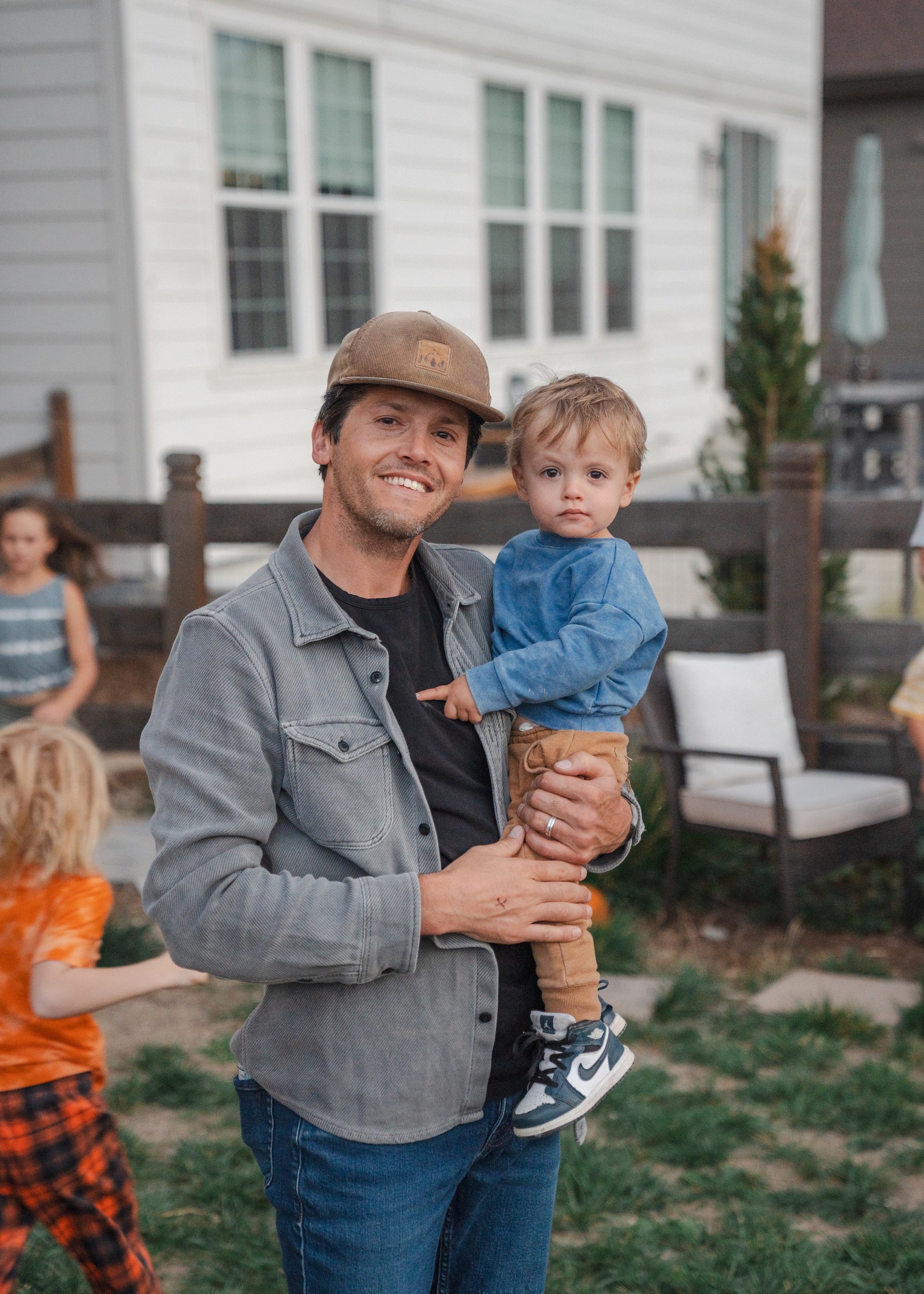 A person in a brimmed hat and gray jacket stands in a yard holding someone in a blue top, with a white house and an outdoor chair behind.
