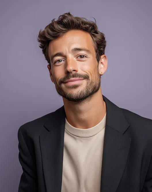 Smiling man with curly brown hair, beard, and a small hoop earring, wearing a beige shirt and black blazer against a purple background.