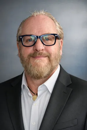 Middle-aged man with blond hair, beard, and glasses wearing a black suit jacket, white shirt, and a necklace with a pendant.