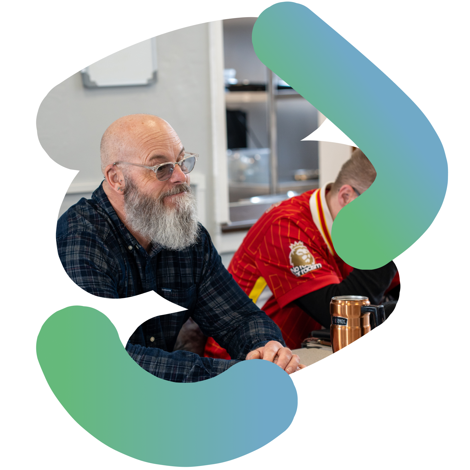 “Older man with a long grey beard and glasses sitting at a table, listening, with another person beside him in a red shirt.