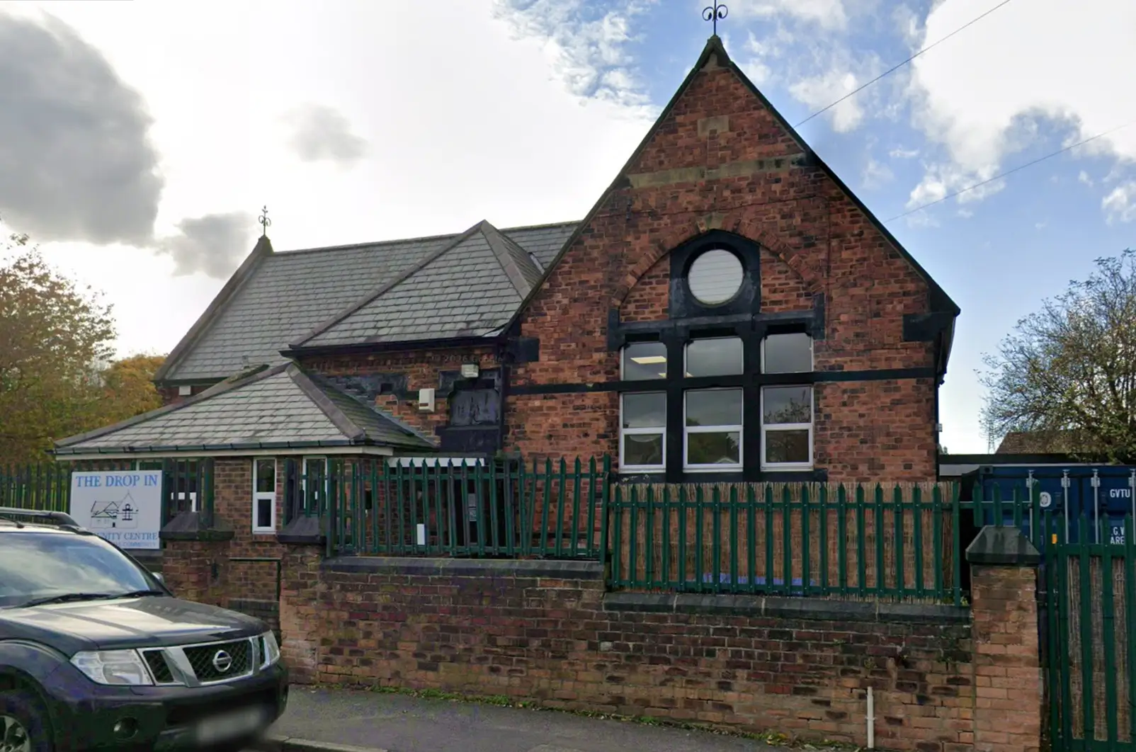 Exterior of The Drop In Centre, a red brick building with a pitched roof, large windows, and a green metal fence along the front.