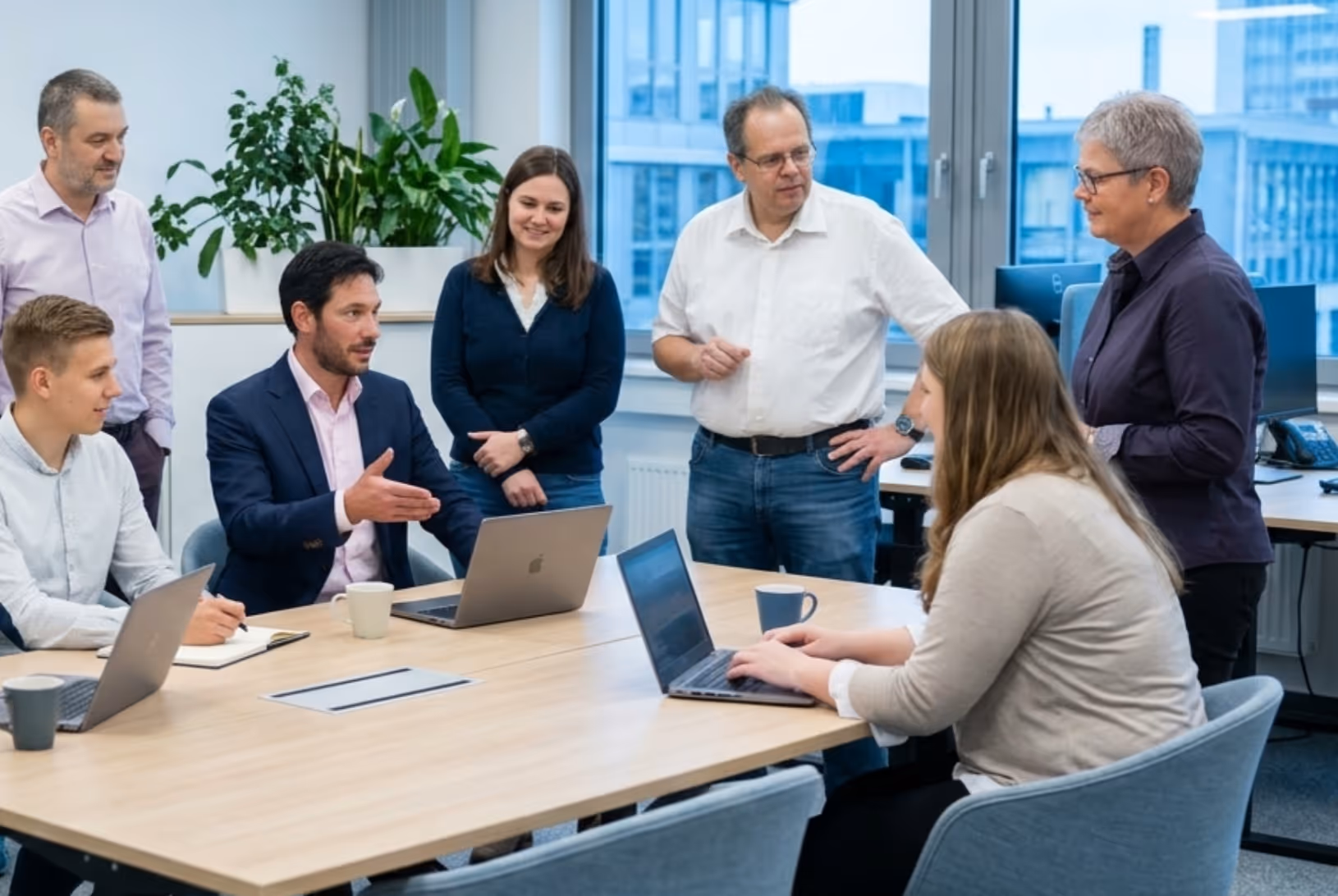 Sechs Personen sprechen in einem modernen Büro bei einem Meeting an einem Tisch mit Laptops.