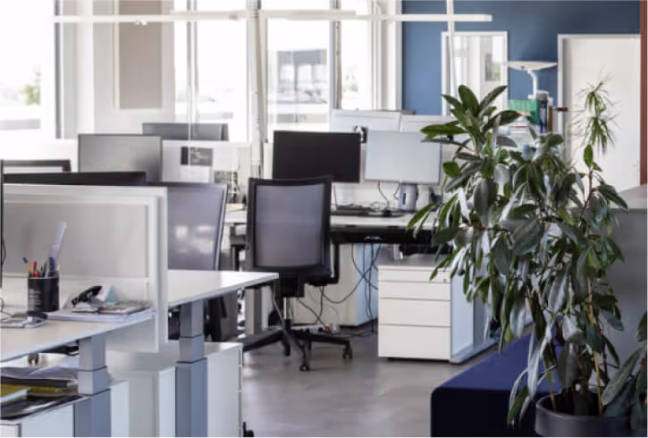 Modernes Büro mit Schreibtischen, Computern und Stühlen in einem hellen Raum mit großen Fenstern.
