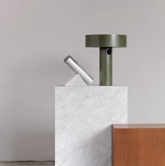 Modern minimalist sculpture with a silver cylindrical piece angled on a white marble triangular base and a green T-shaped structure on a white marble pedestal beside a ribbed brown rectangular object.