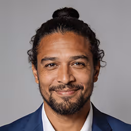 Smiling man with curly hair tied back and facial hair wearing a dark shirt.