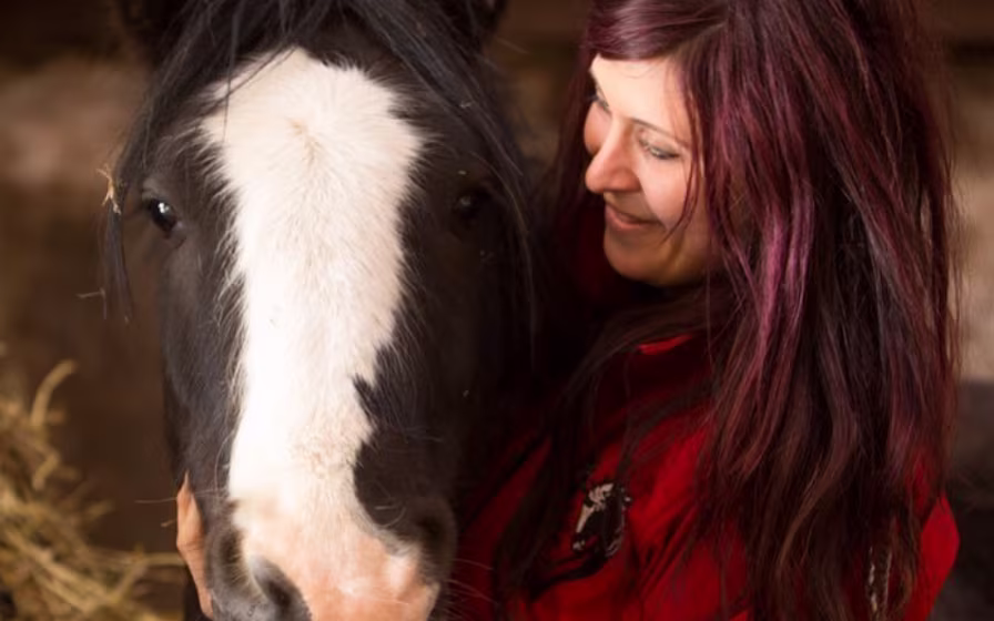 Woman with long dark hair smiling and hugging a black and white horse inside a barn.