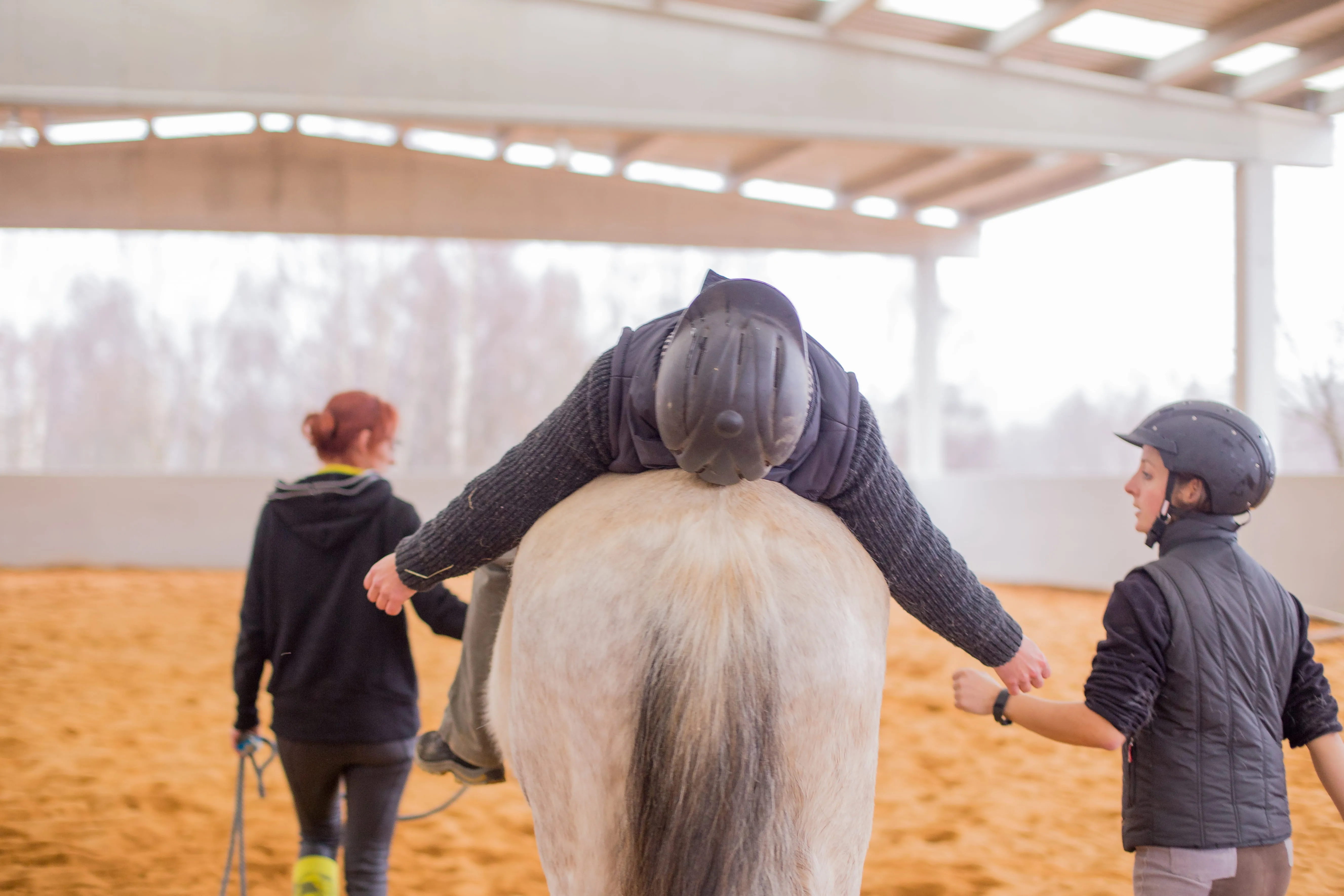 Therapist assists child lying on a horse during a hippotherapy session indoors.