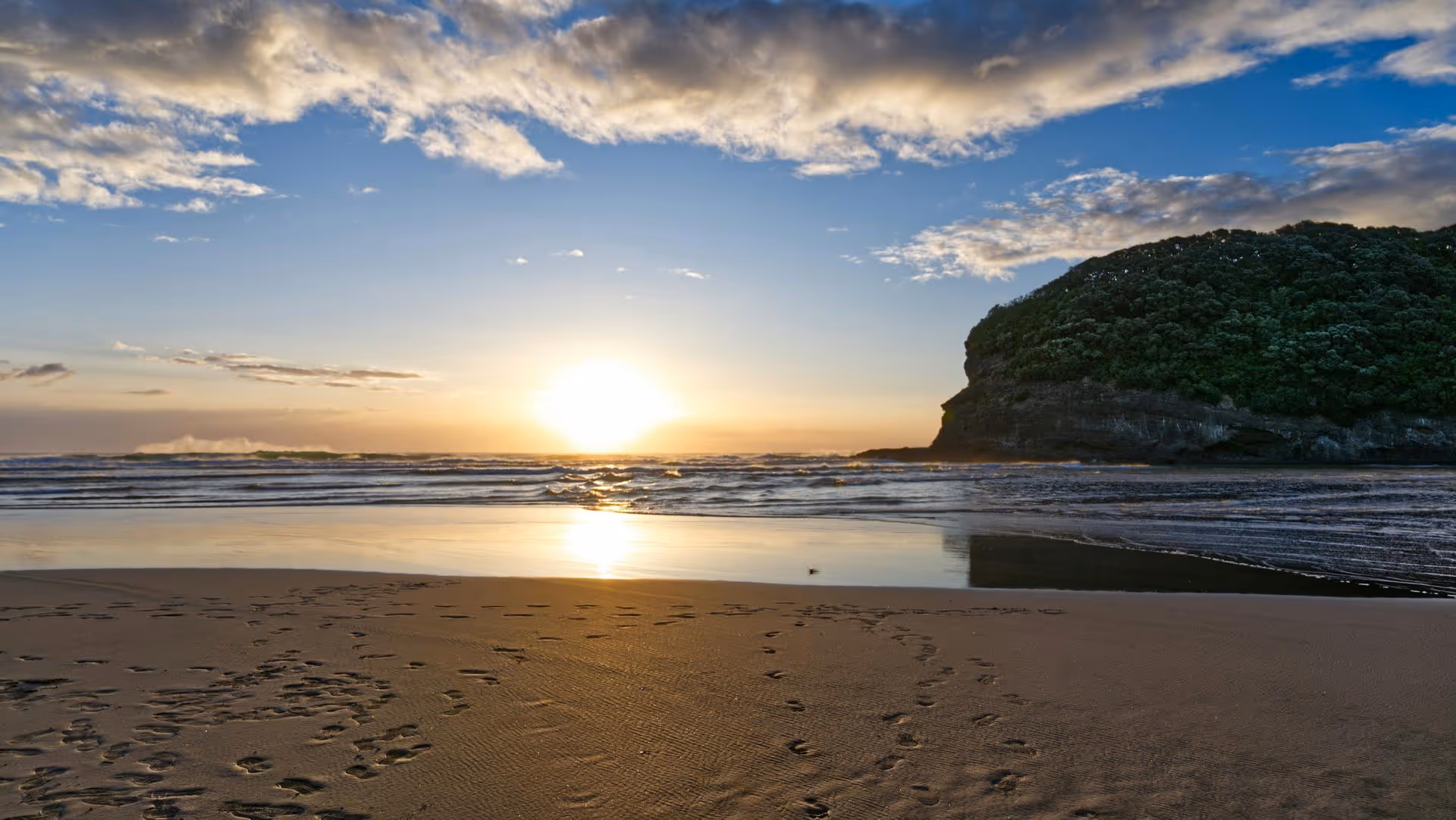 Sunset over a beach with calm waves, wet sand with footprints, and a green cliff on the right.