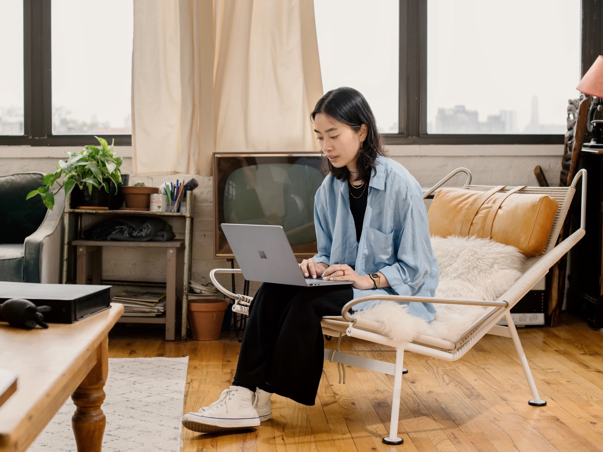 Young woman sitting on a modern chair with a fur throw, using a laptop in a sunlit living room with wooden floors and large windows.