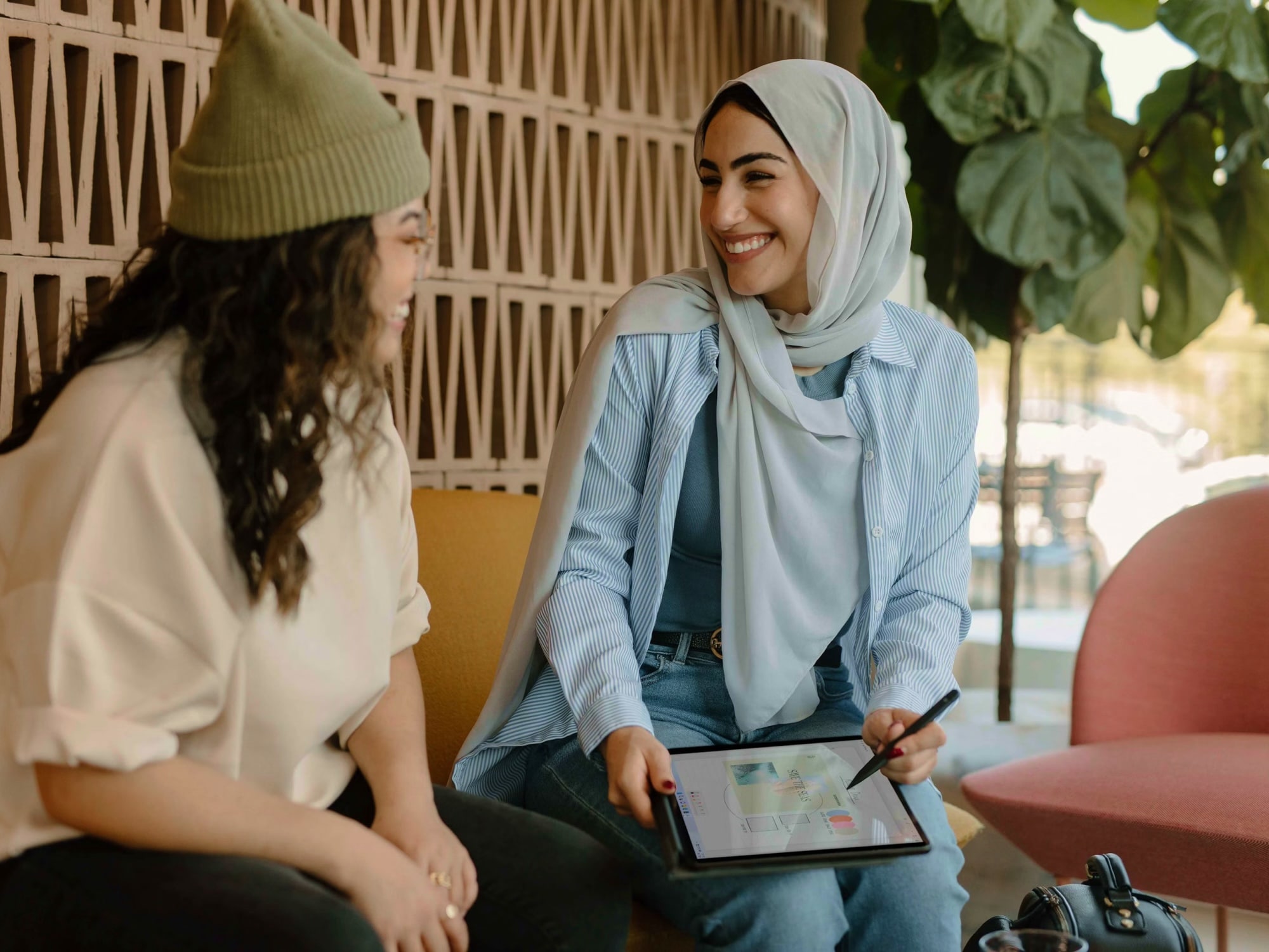 Two women seated and smiling, one wearing a hijab and holding a tablet with a stylus showing a colorful digital design.