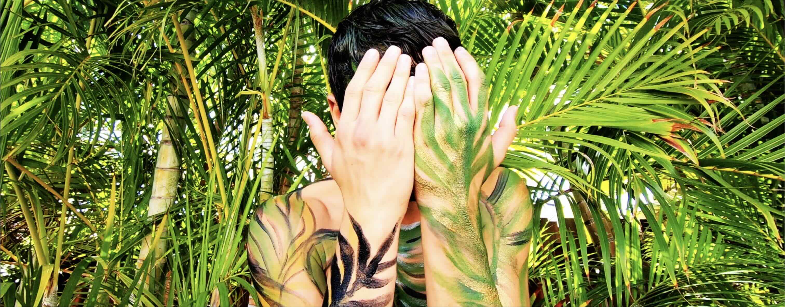 Still image from music video boy painted in green and black blending in with palm plants behind him