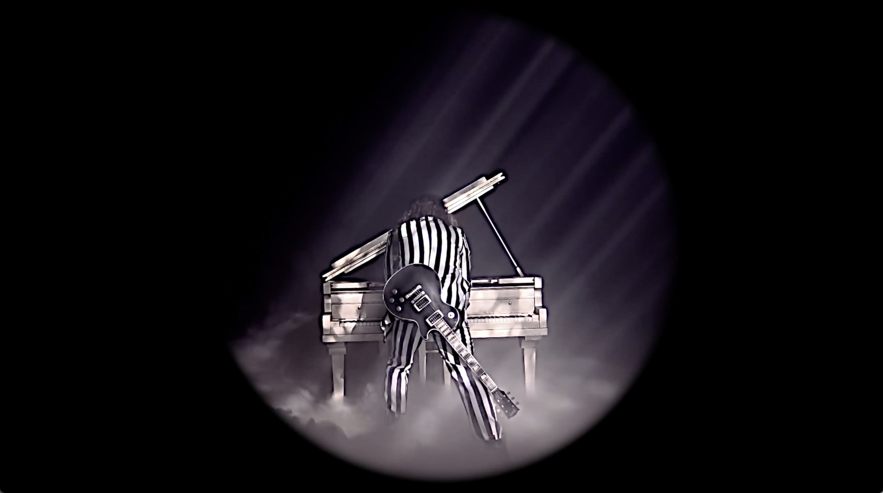 Music video still image man wearing black and white suite playing piano with guitar strapped to his back