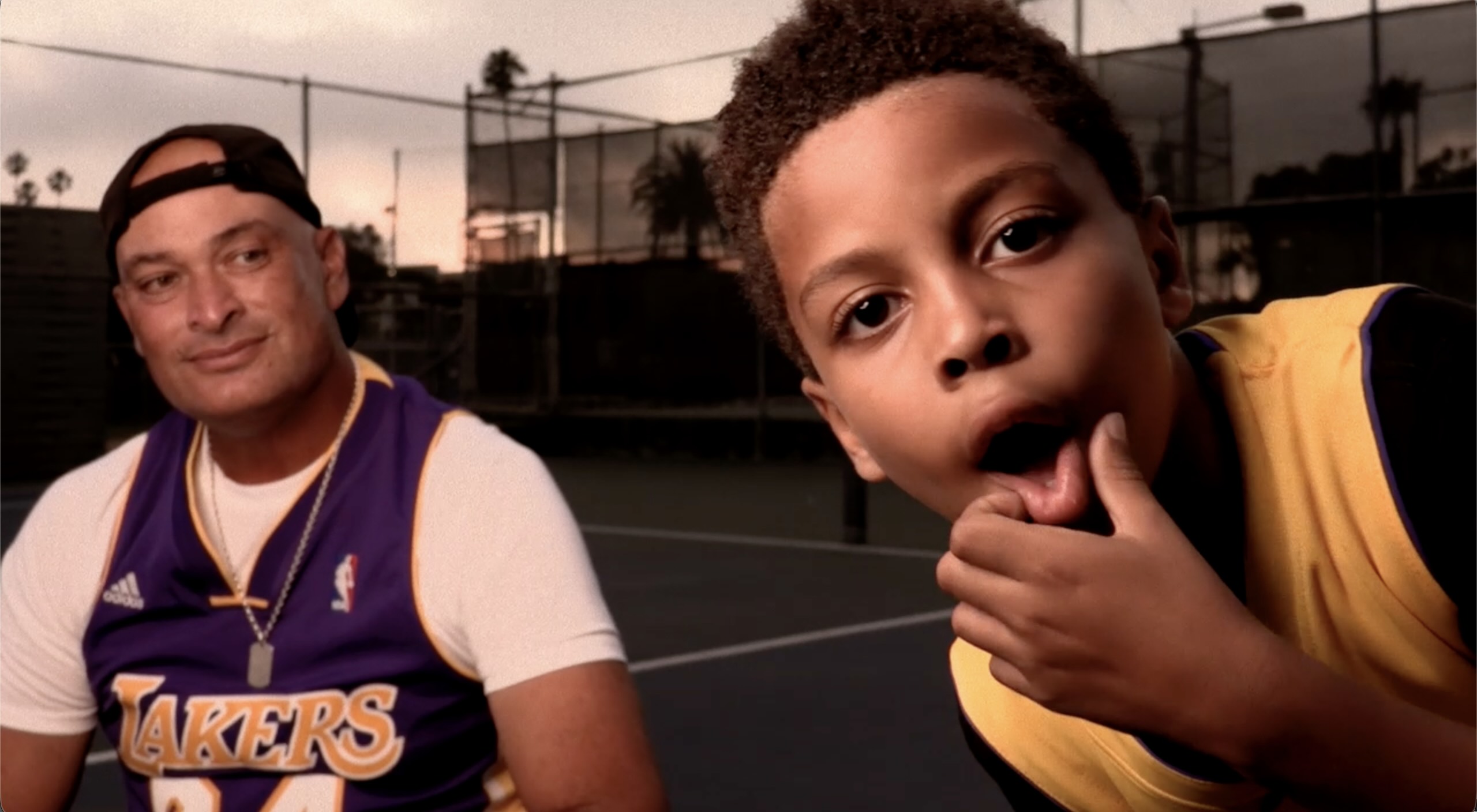 Still image from music video young boy staring into camera with man behind him on basketball court