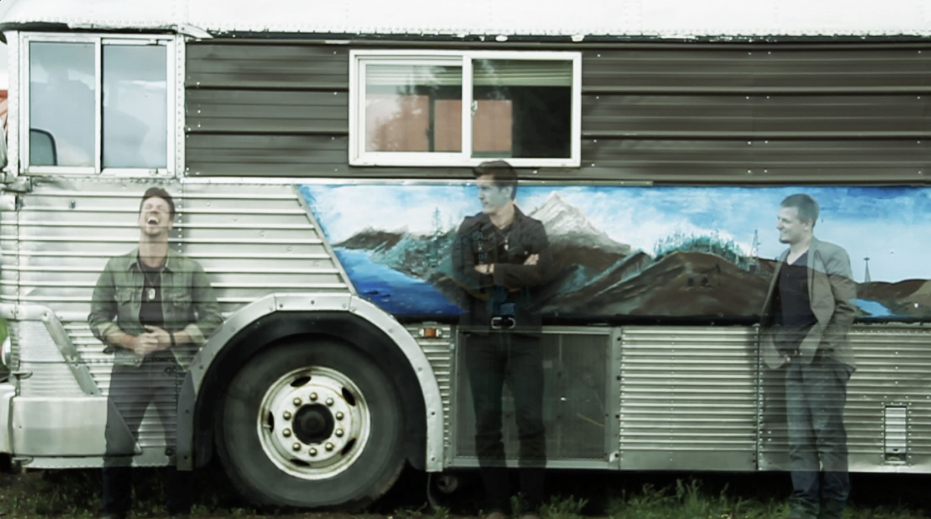Three men standing in mid laugh against tour bus with a mountainside painted on the side of the bus still from music video