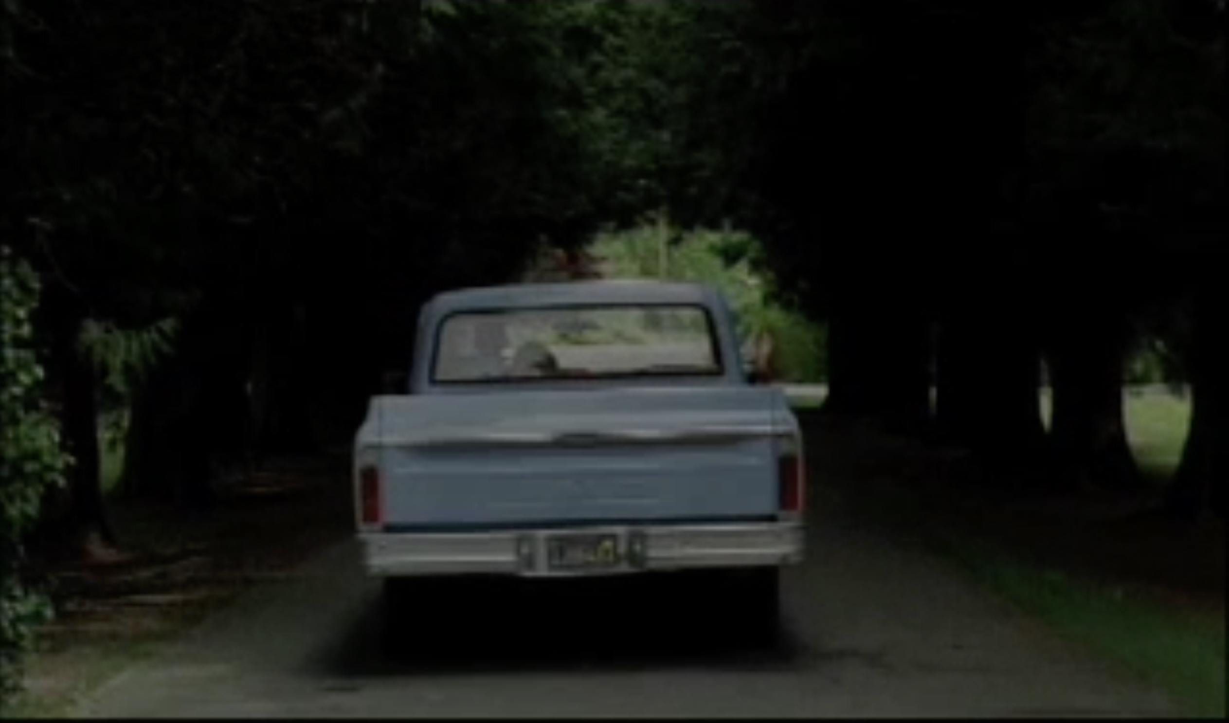 Still image from country music video old truck driving between canopy of trees man driving woman leaned against him with her feet hanging outside her passenger window