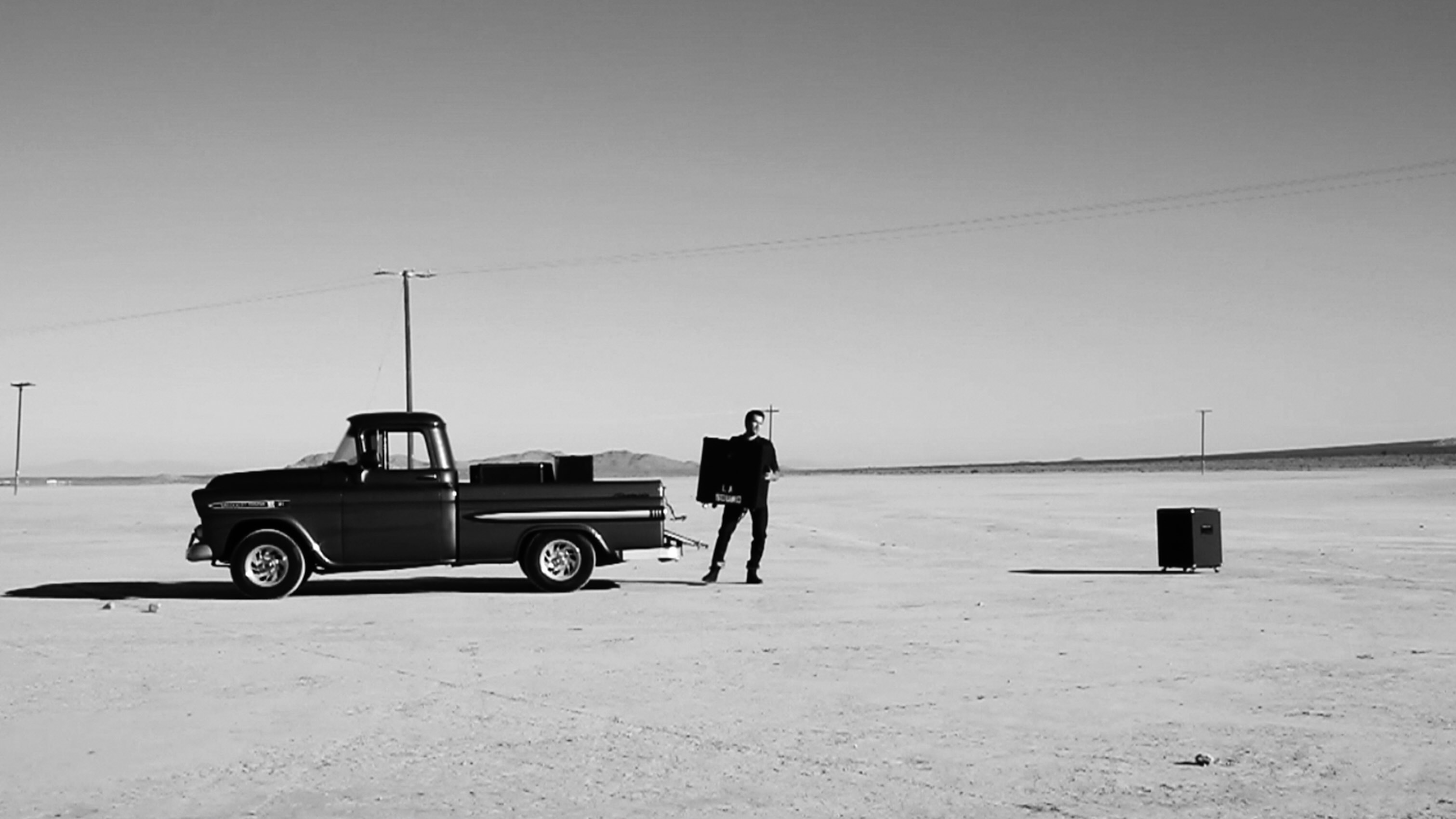 Black and white wide shot of man carrying large speaker from vintage truck bed with many speakers and placing them in the desert still image from music video